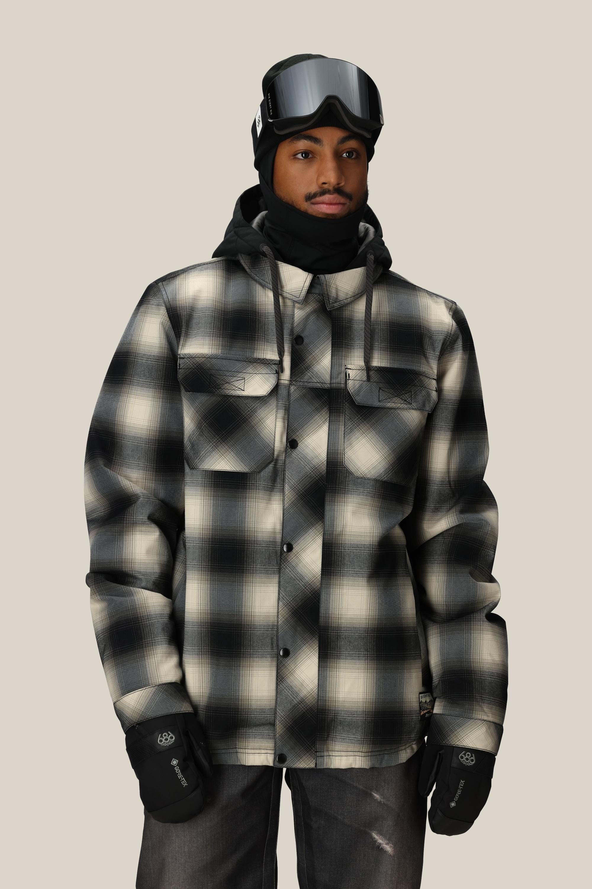 A person wearing the 686 Men's Woodland™ Insulated Jacket stands against a plain, light background, paired with black gloves, a black hood, neck warmer, and ski goggles.