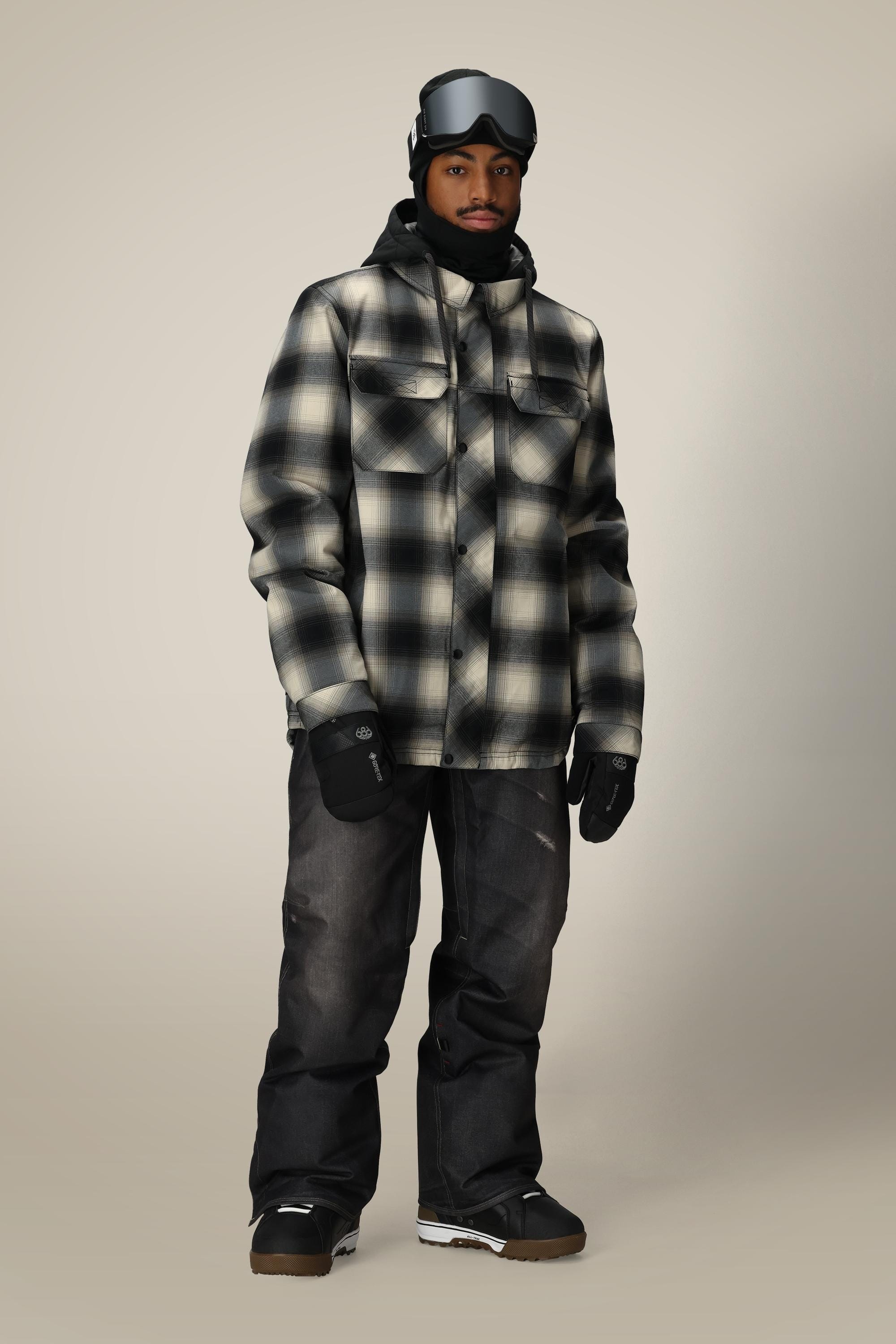A person wears a 686 Men's Woodland™ Insulated Jacket by 686 in black and white plaid, paired with black gloves, dark snow pants, brown boots, and black ski goggles, standing facing forward against a plain background.