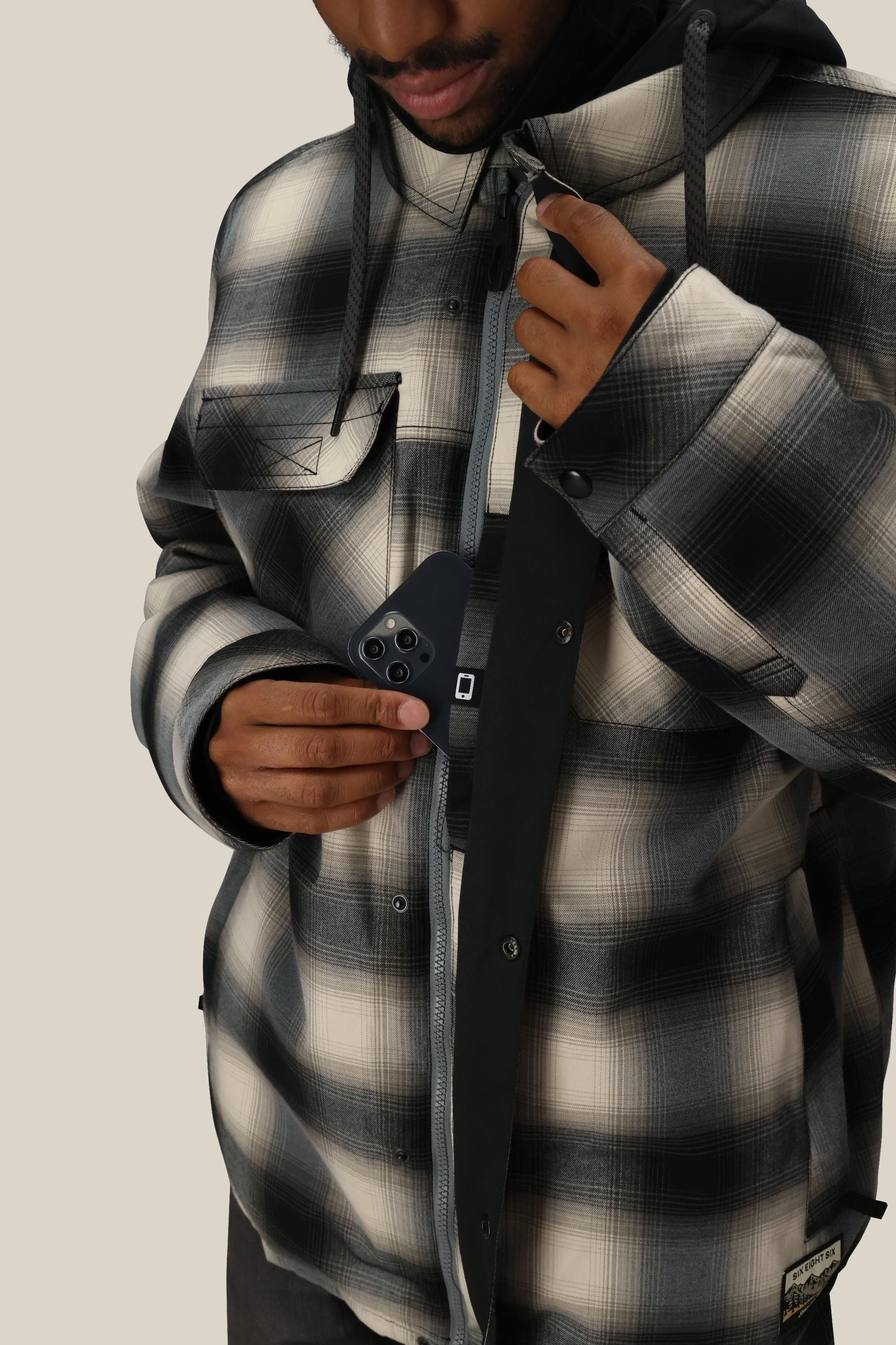 A person wearing the 686 Men's Woodland™ Insulated Jacket unzips the black-and-white plaid jacket to place a black smartphone with three rear cameras into an inside pocket, with the image cropped at the mouth to showcase the clothing and action.