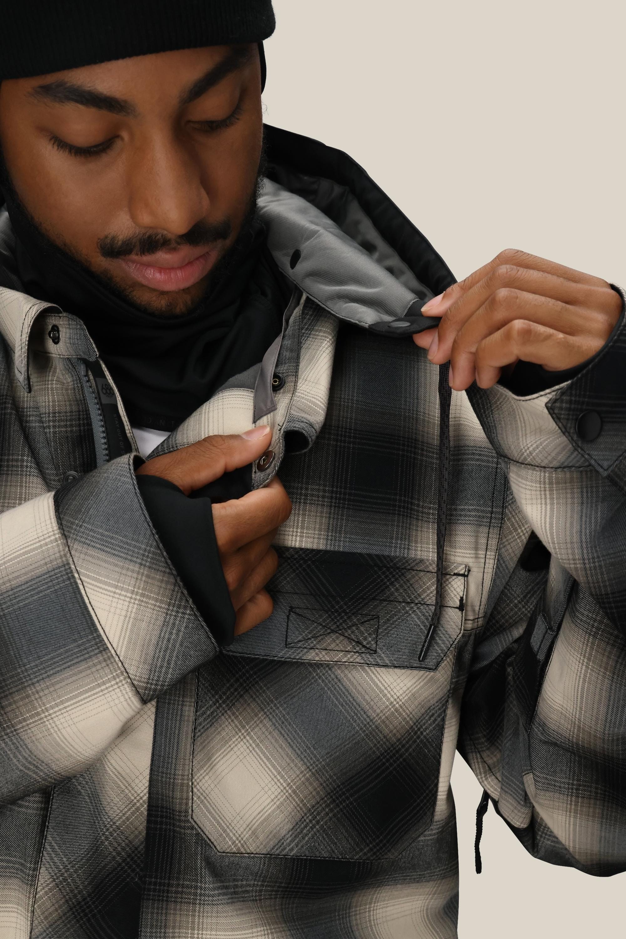 A man wears the 686 Men's Woodland™ Insulated Jacket by 686 in black, gray, and white plaid, paired with a black beanie. He fastens the snap buttons near the collar while looking down with a slight smile.