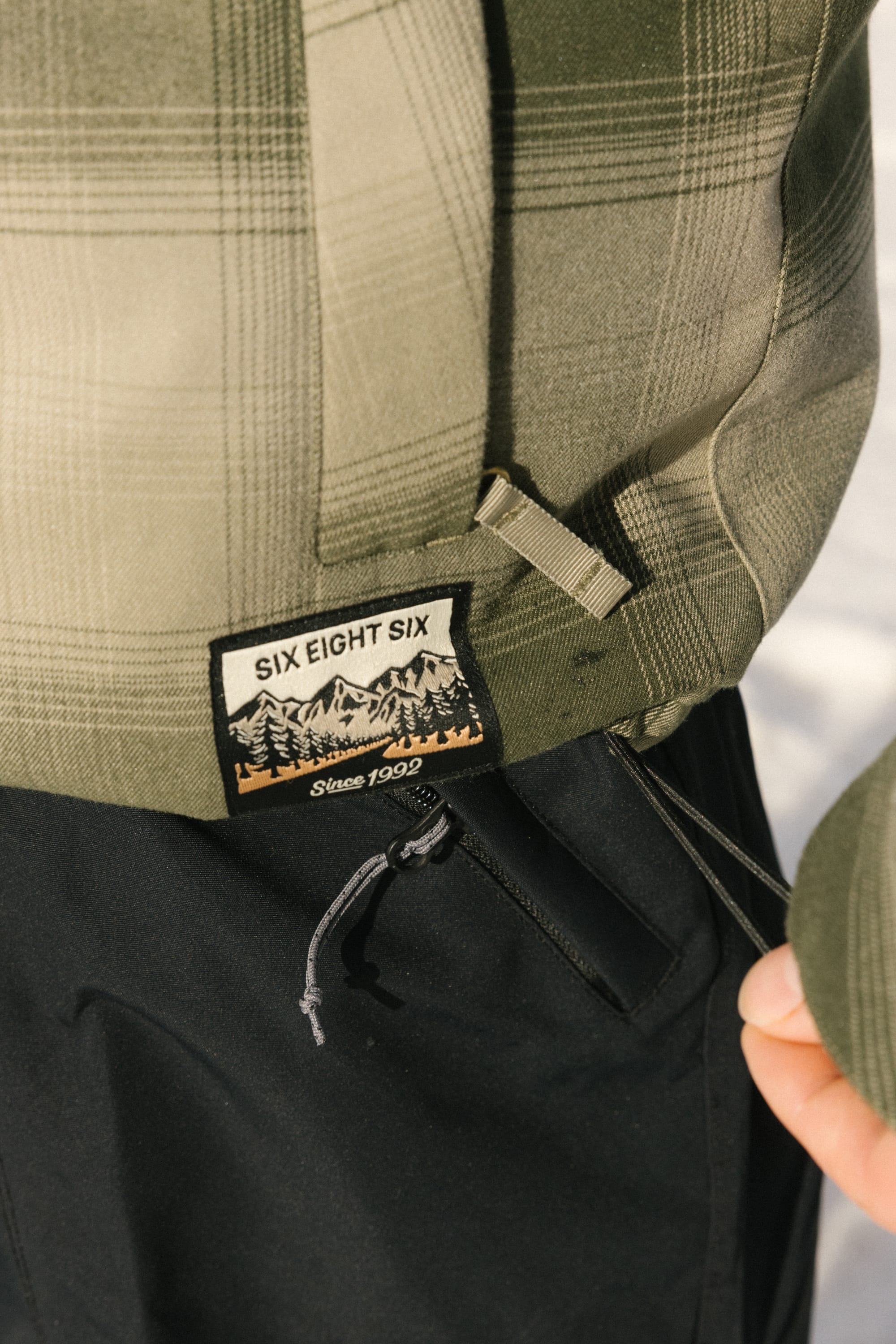 Close-up of the 686 Men's Woodland™ Insulated Jacket in green plaid with a woven "686 Since 1992" label above a mountain and tree design. The wearer pairs it with black pants, holding the jacket’s hem.