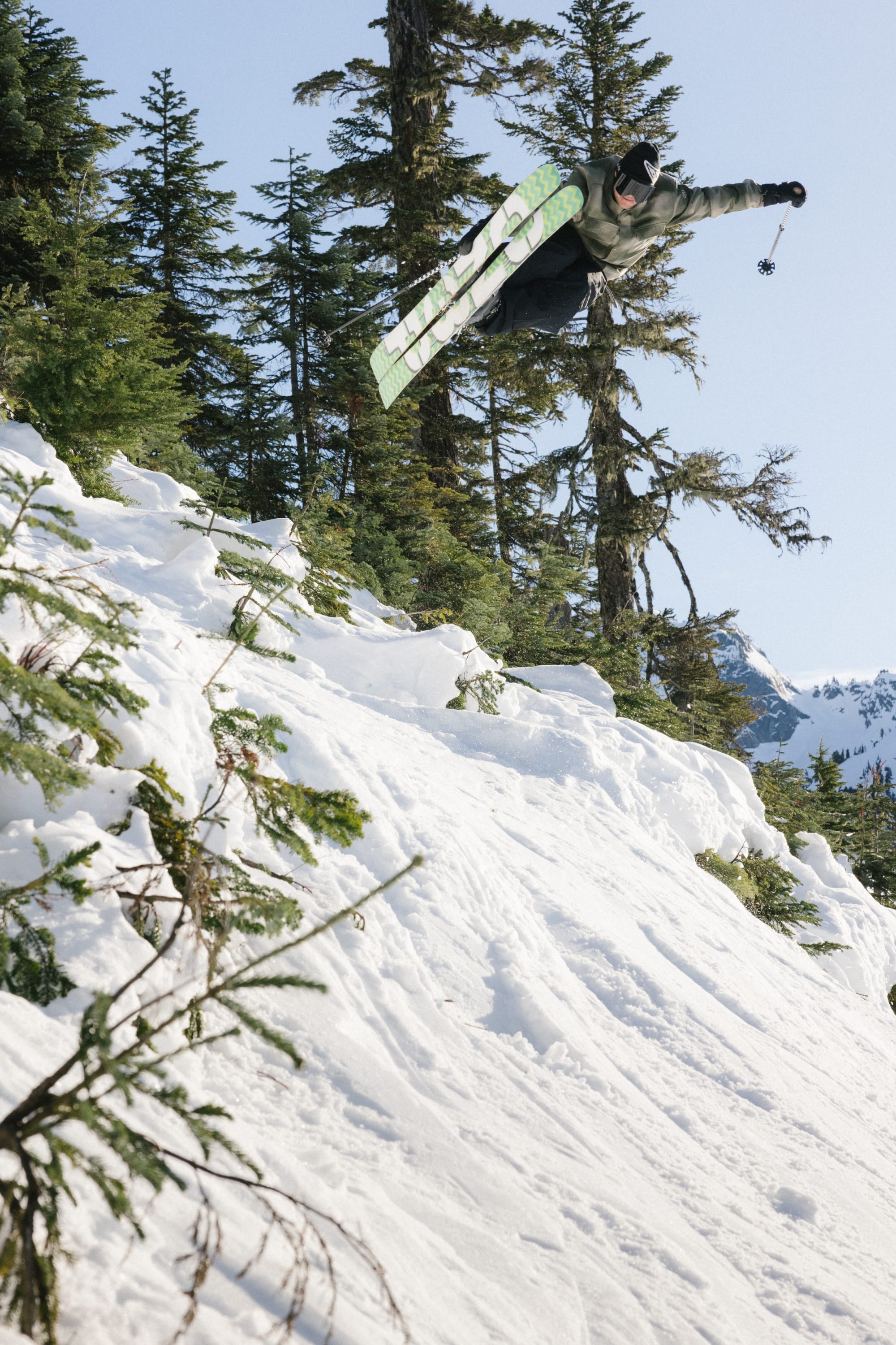 A snowboarder catches air off a snowy slope, wearing the 686 Men's Woodland™ Insulated Jacket by 686, surrounded by evergreen trees and clear blue sky, capturing the excitement of snowboarding.