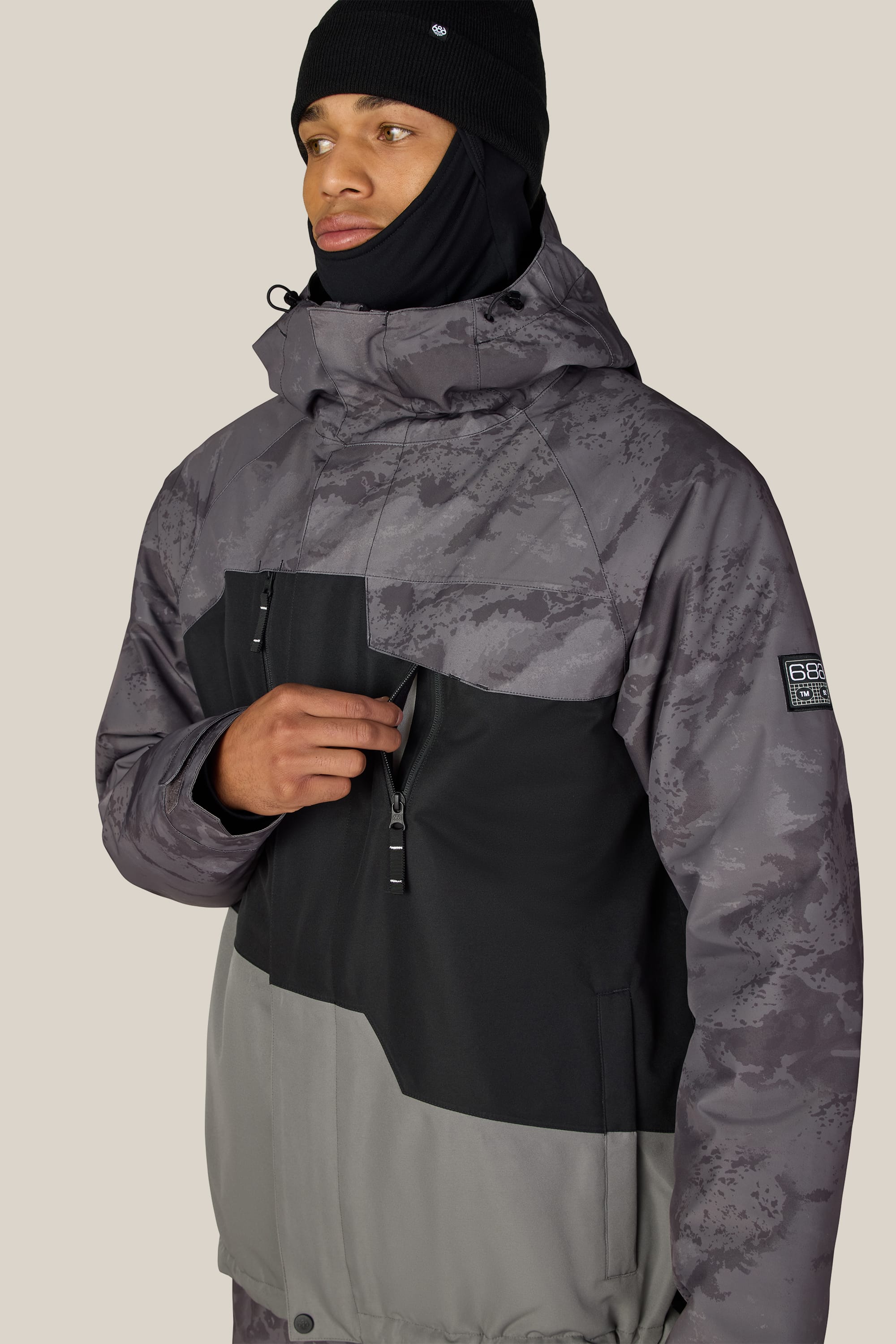 A person wears the 686 Men’s Geo™ Insulated Jacket by 686—a gray and black men’s waterproof jacket with a hood—along with a black beanie and black face covering, standing against a plain beige background while holding the front zipper.