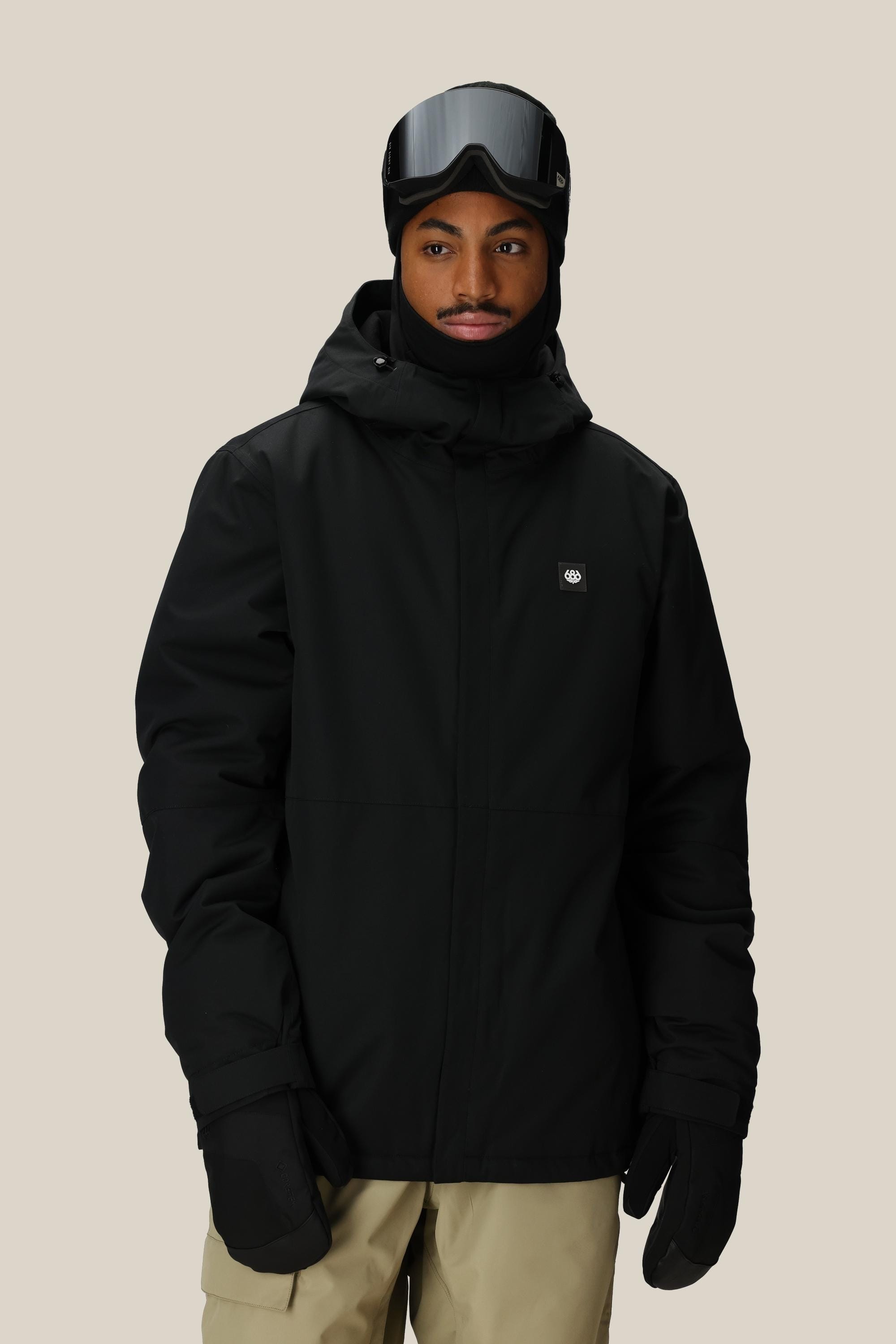 A person wearing the 686 Men's Foundation™ Insulated Jacket with a hood, along with black gloves, balaclava, and ski goggles, stands against a beige background. They have a short beard and look slightly to the side.