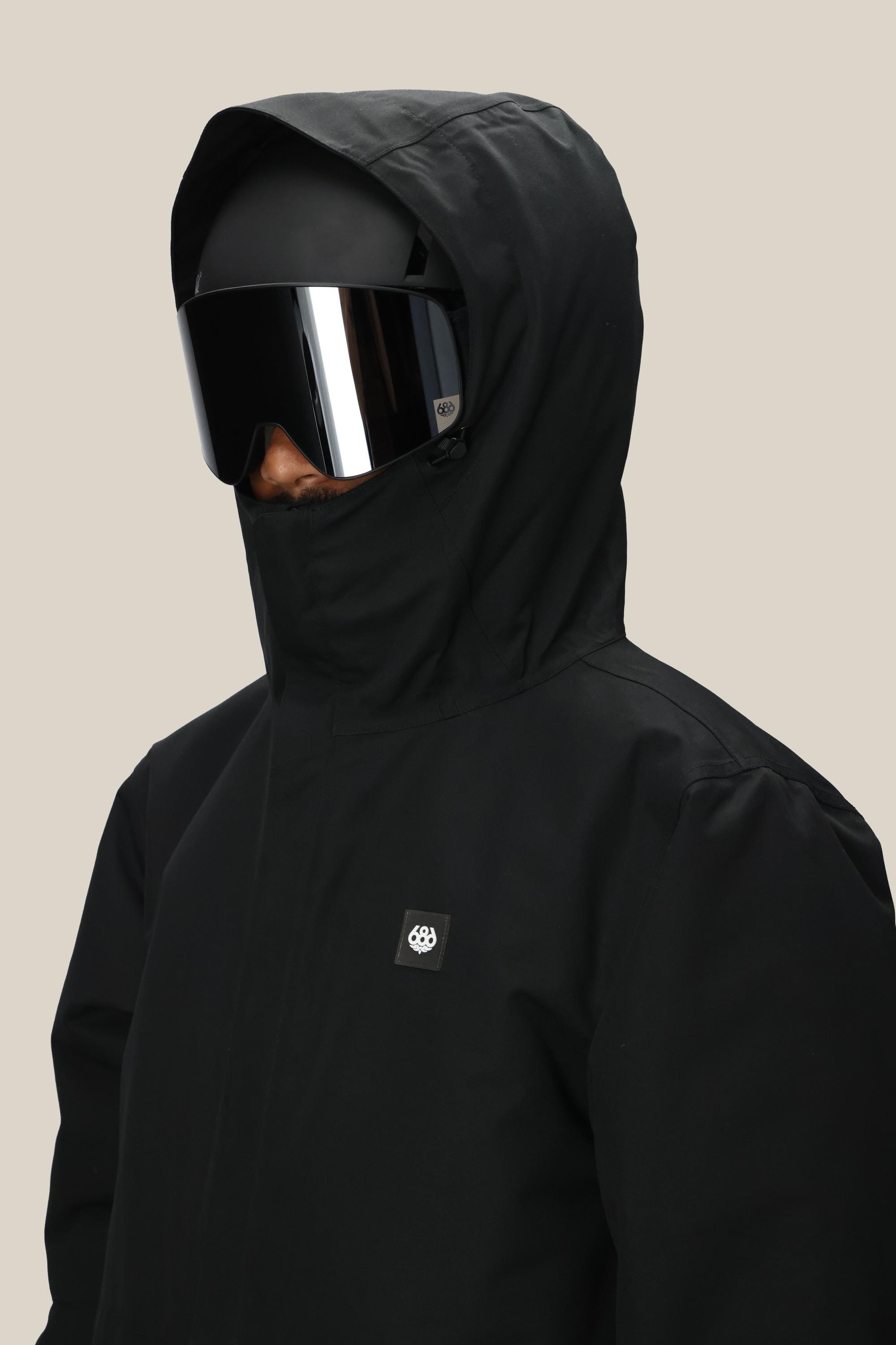 A person wearing the 686 Men’s Foundation™ Insulated Jacket in black, paired with a black helmet and large reflective ski goggles, is facing slightly to the side against a plain beige background.