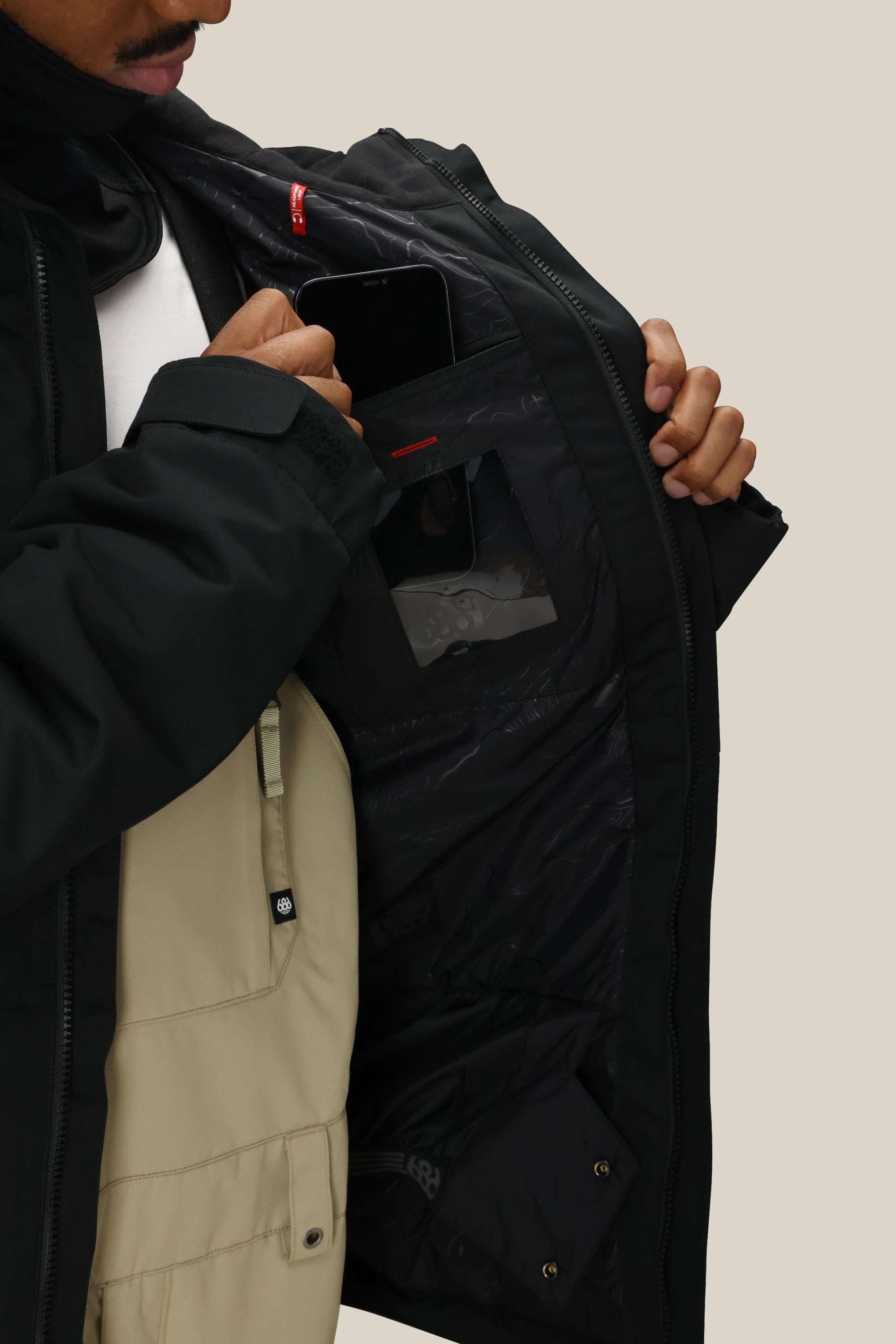 A person opens the 686 Men's Foundation™ Insulated Jacket to show inner pockets—one holding a black smartphone—with a beige layer worn underneath.