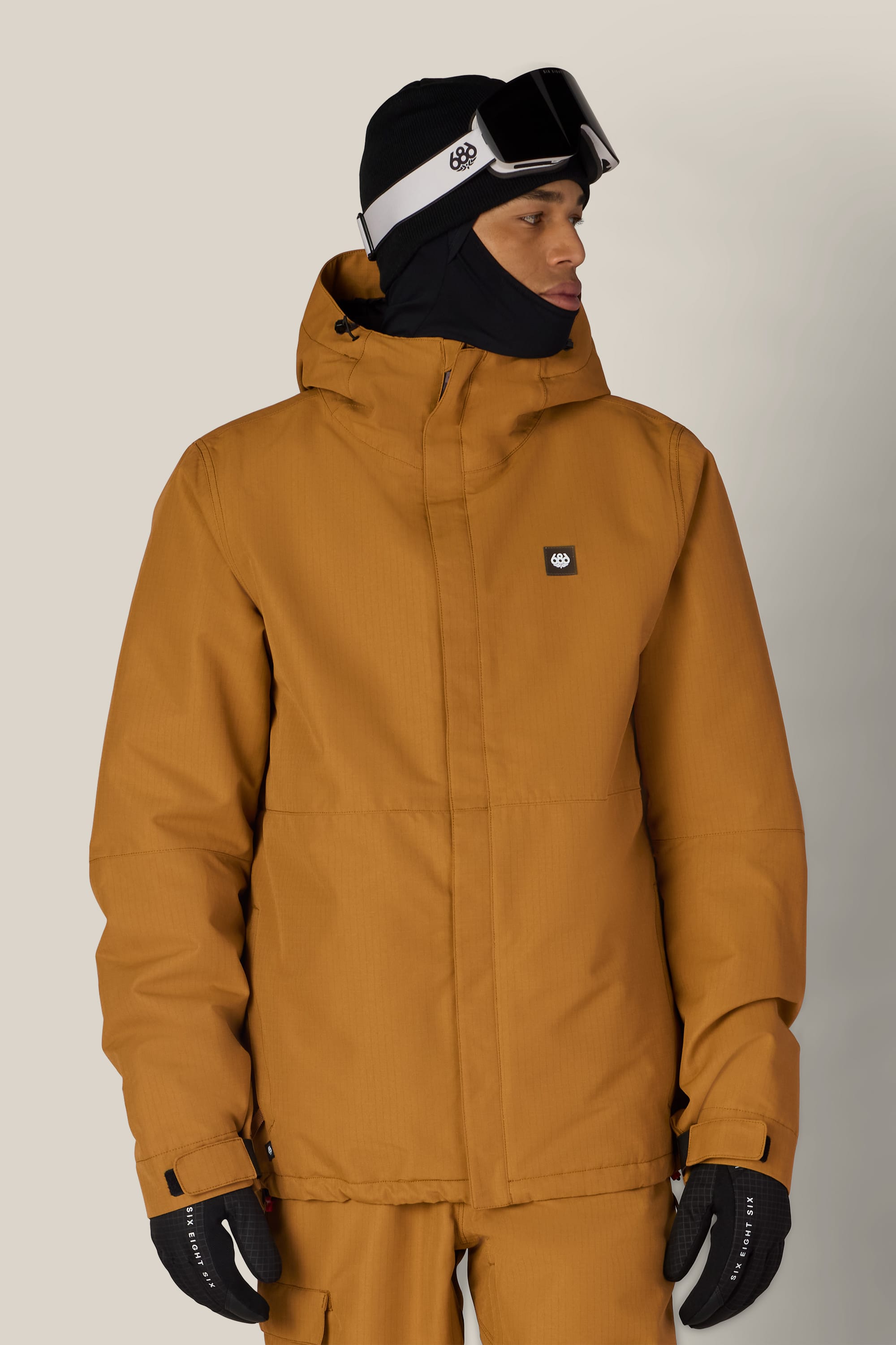 A person wearing the 686 Men’s Foundation™ Insulated Jacket in mustard-yellow, paired with matching pants, black gloves, a black beanie, and ski goggles looks to the side against a plain background.