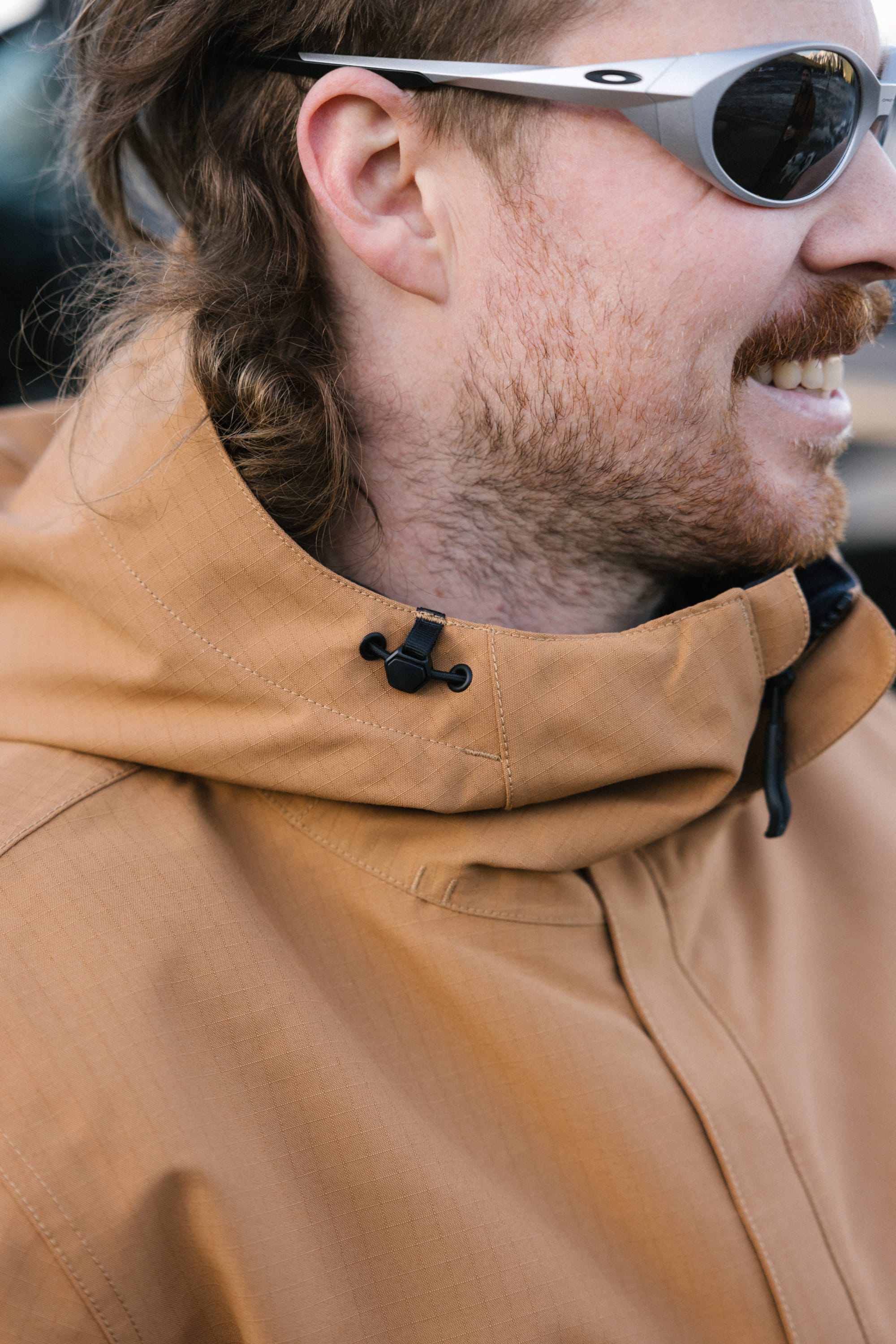 A close-up of a man with light facial hair, wearing silver sunglasses and the 686 Men's Foundation™ Insulated Jacket with a hood, smiling and looking to the right.