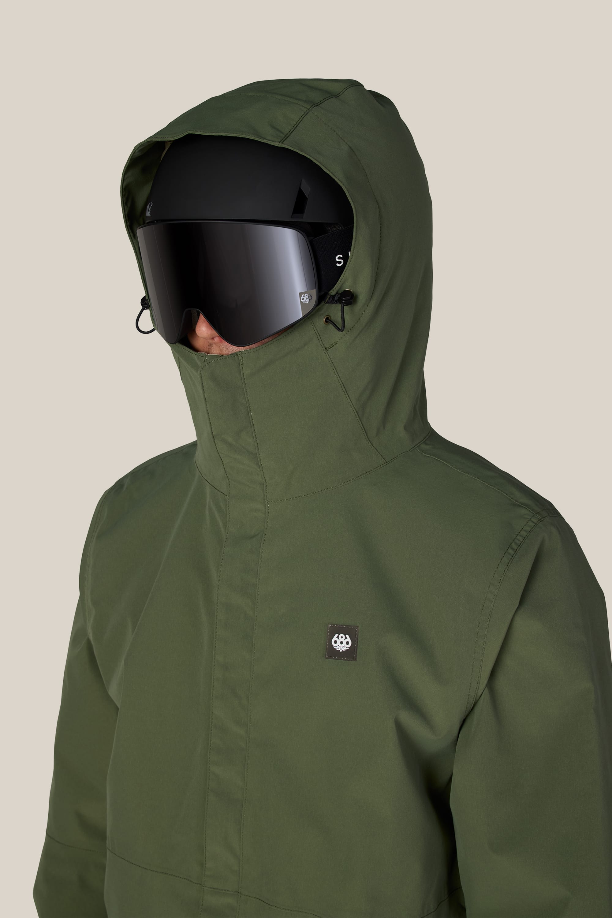 A person wearing the 686 Men’s Foundation™ Insulated Jacket in olive green, featuring a small chest logo patch, and black ski goggles faces forward against a plain beige background.