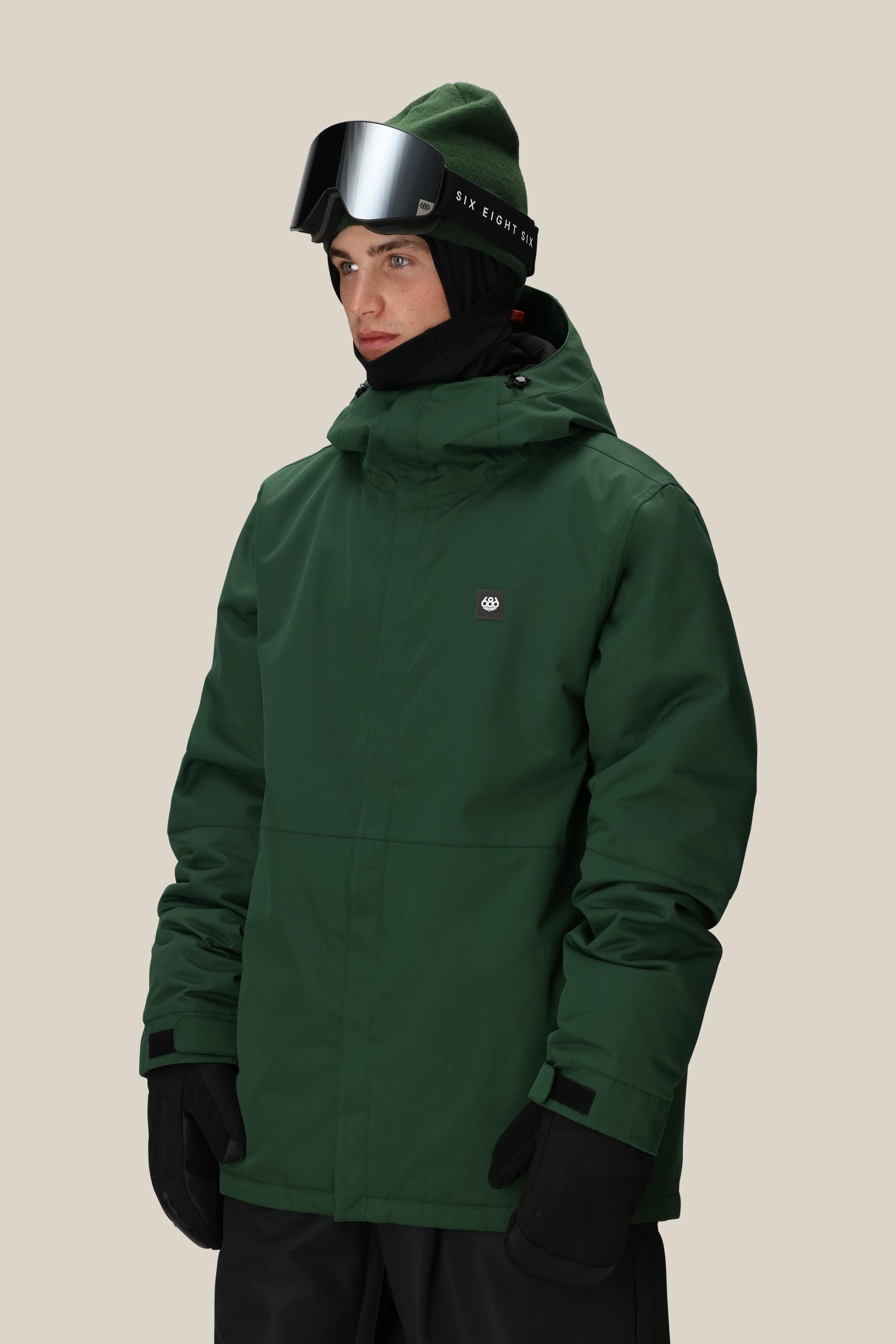 A man wearing the 686 Men's Foundation™ Insulated Jacket in dark green, a green beanie, black gloves, and ski goggles stands against a plain beige background.