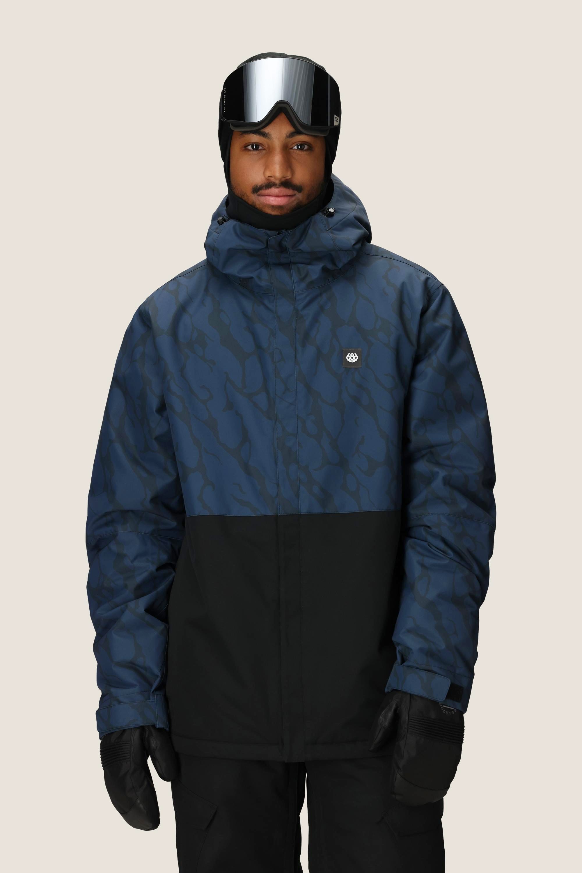 A person in a 686 Men's Foundation™ Insulated Jacket by 686, black gloves, ski goggles, and a black beanie stands against a plain light background.