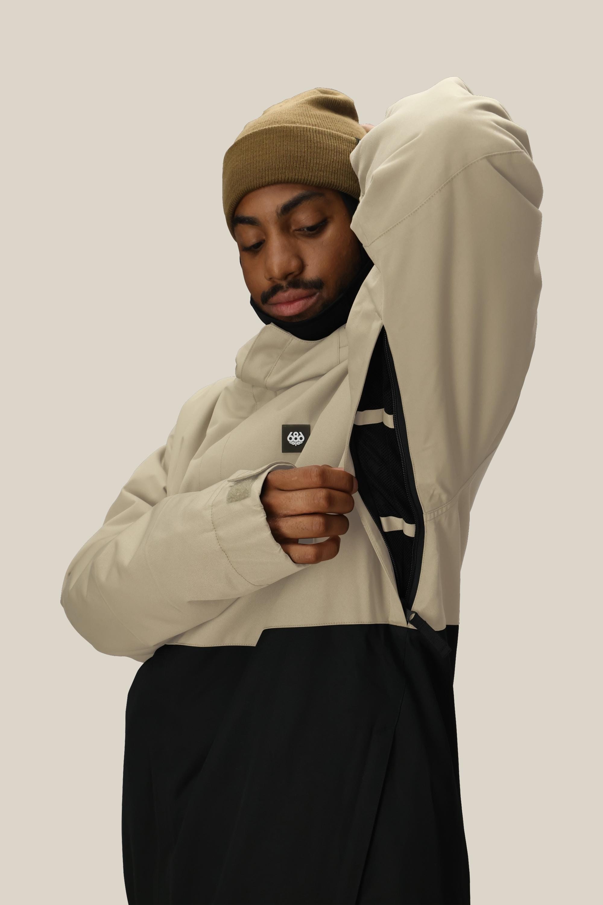 A person wearing a beige beanie and the 686 Men's Foundation™ Insulated Jacket in beige-and-black by 686 unzips a side zipper to show the jacket's inner lining, set against a plain light beige background.