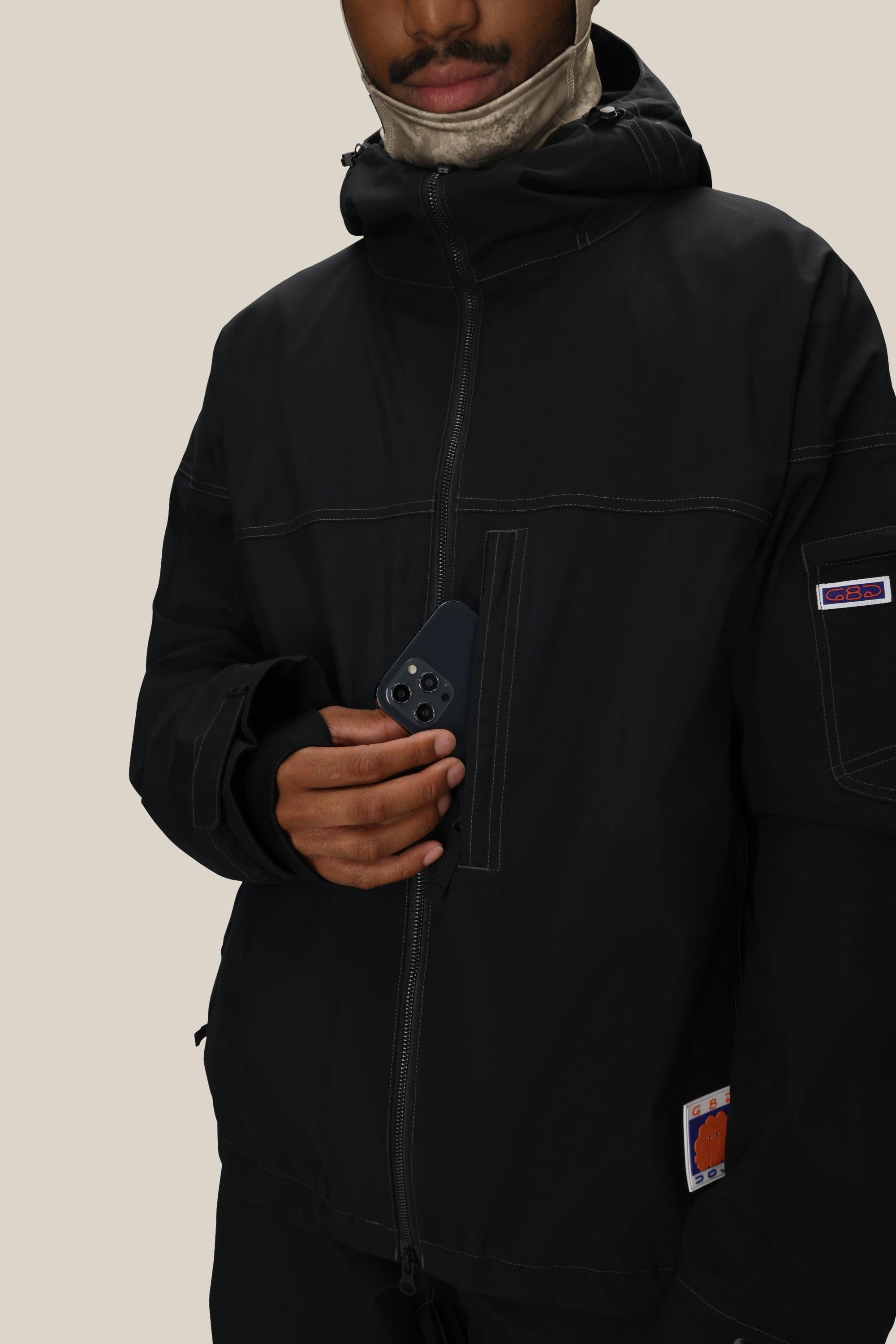A person in a black 686 Dojo® Jacket with a hood holds a smartphone featuring a triple camera. Only the lower face is visible, and the background is plain beige.