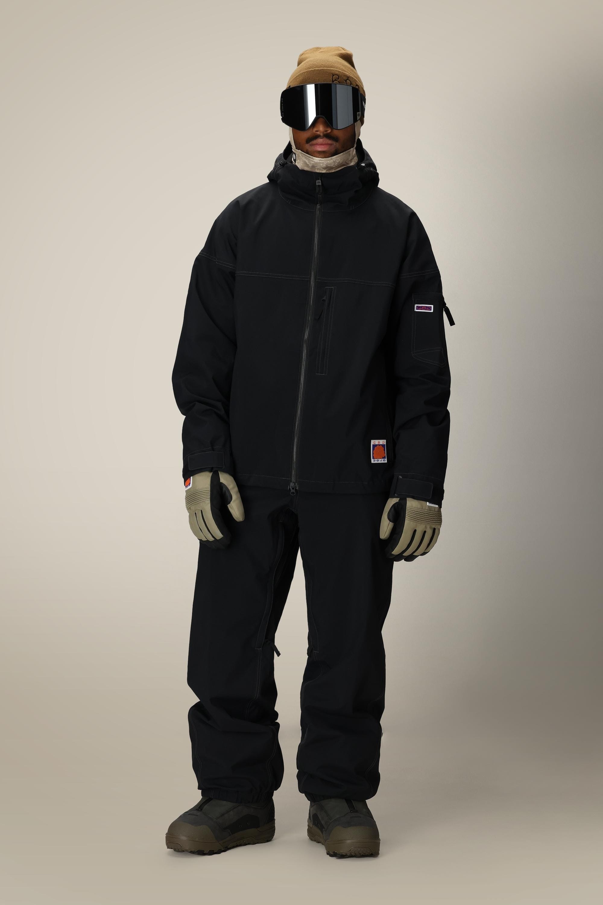 A person stands facing forward wearing a black 686 Dojo® Jacket by 686, matching nylon waterproof pants, beige gloves, brown boots, a tan beanie, and large reflective ski goggles against a plain background.