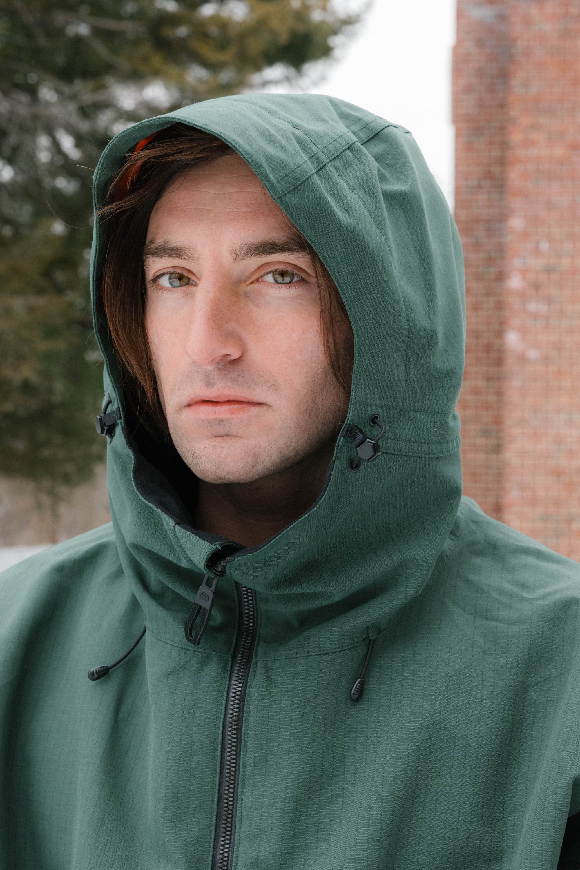 A person with long brown hair wears the 686 Dojo® Jacket by 686, featuring a green hood, while standing outdoors on a cloudy day. Trees and a brick wall appear in the background as they face the camera with a neutral expression.