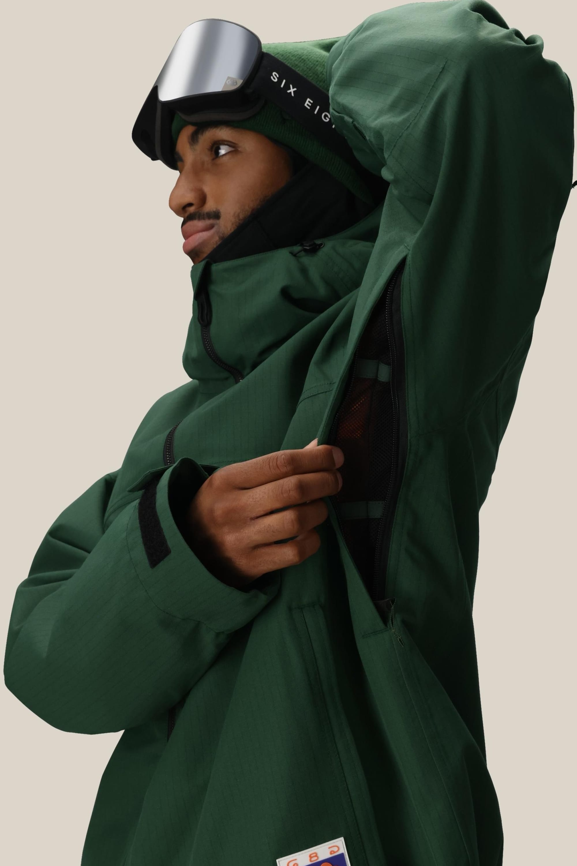 A person in a green 686 Dojo® Jacket and beanie unzips the underarm vent. They have ski goggles on their head and gaze to the side against a neutral background.