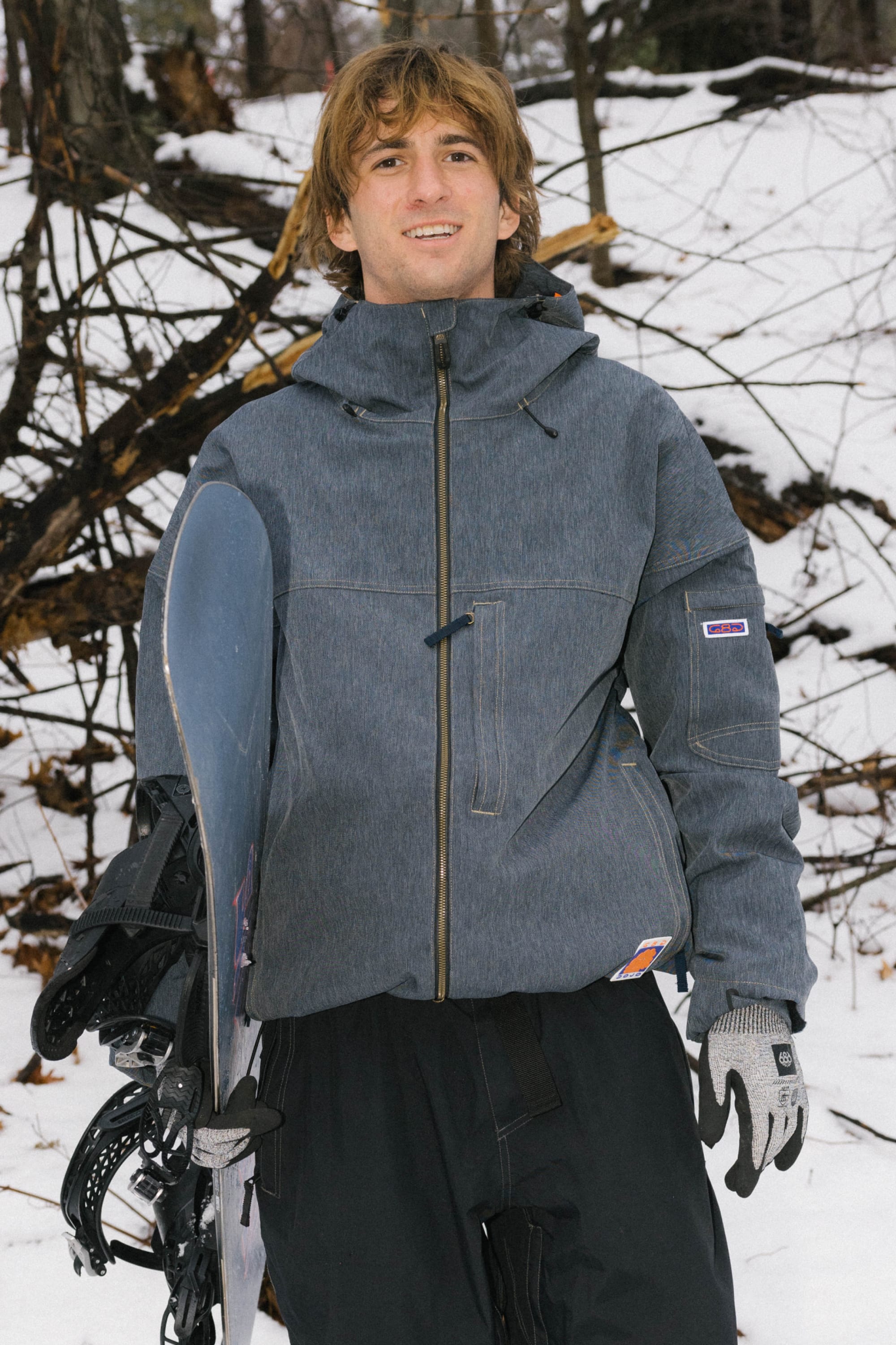 A person with light brown hair, dressed in the 686 Dojo® Jacket and black pants with gloves, stands outdoors in the snow holding a snowboard. Bare tree branches and snow are visible in the background.