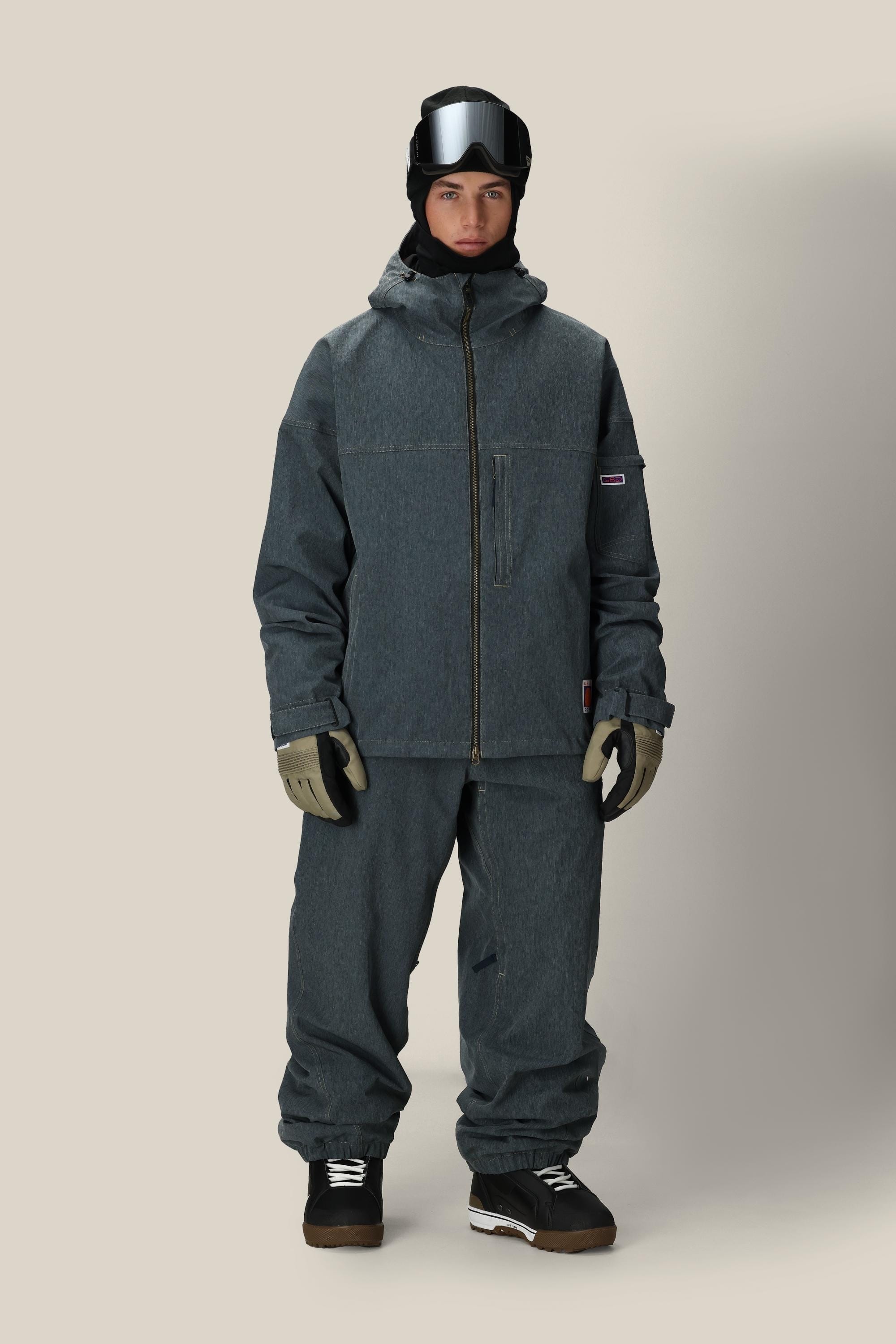 A person faces forward in a gray 686 Dojo® Jacket by 686, matching loose pants, gloves, black ski goggles, a black hat, and black sneakers with white soles against a plain beige background.