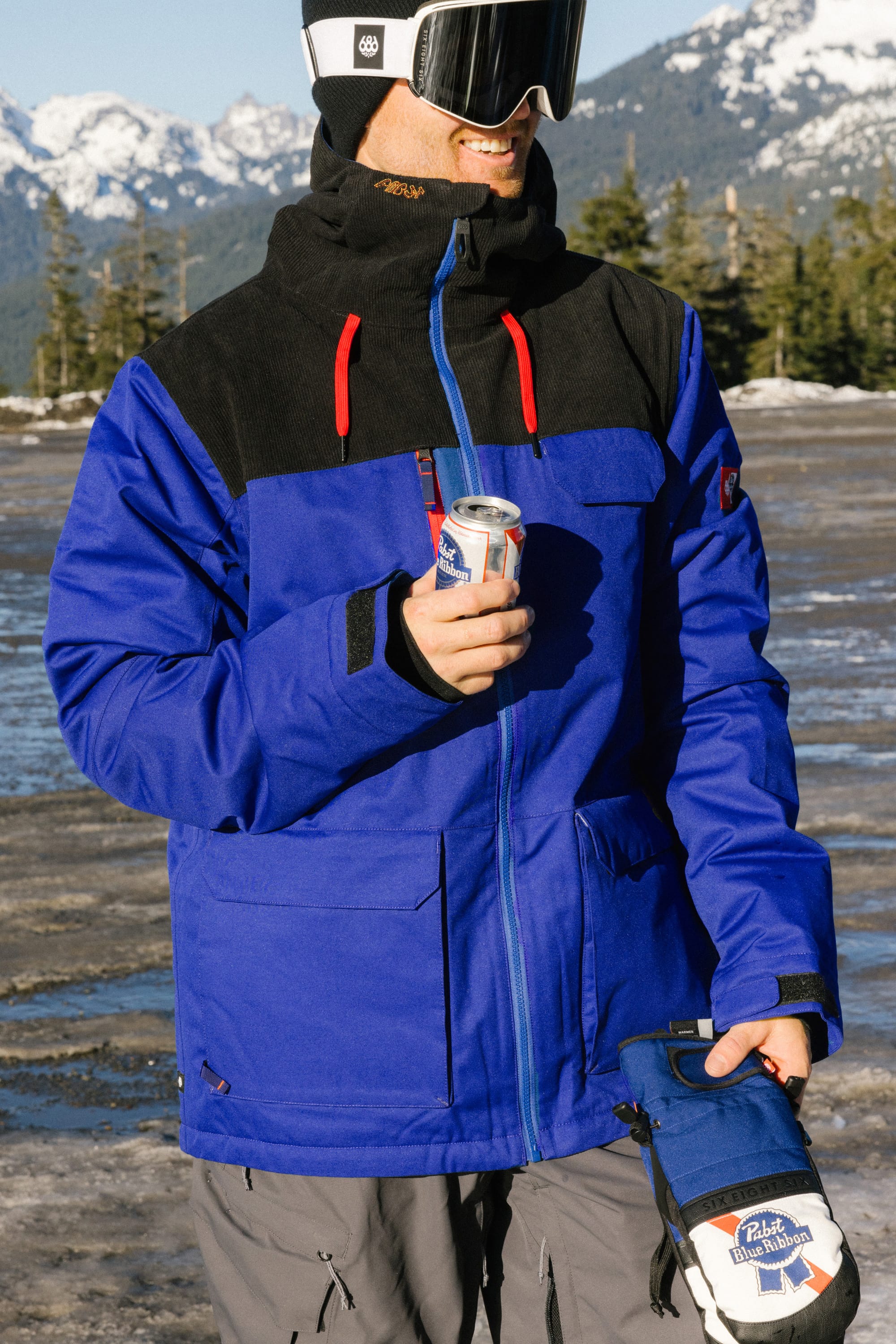 Wearing the 686 Mens PBR Cooler Insulated Jacket, a person with ski goggles stands outdoors on a snowy day holding a can of Pabst Blue Ribbon, with pine trees and mountains in the background.