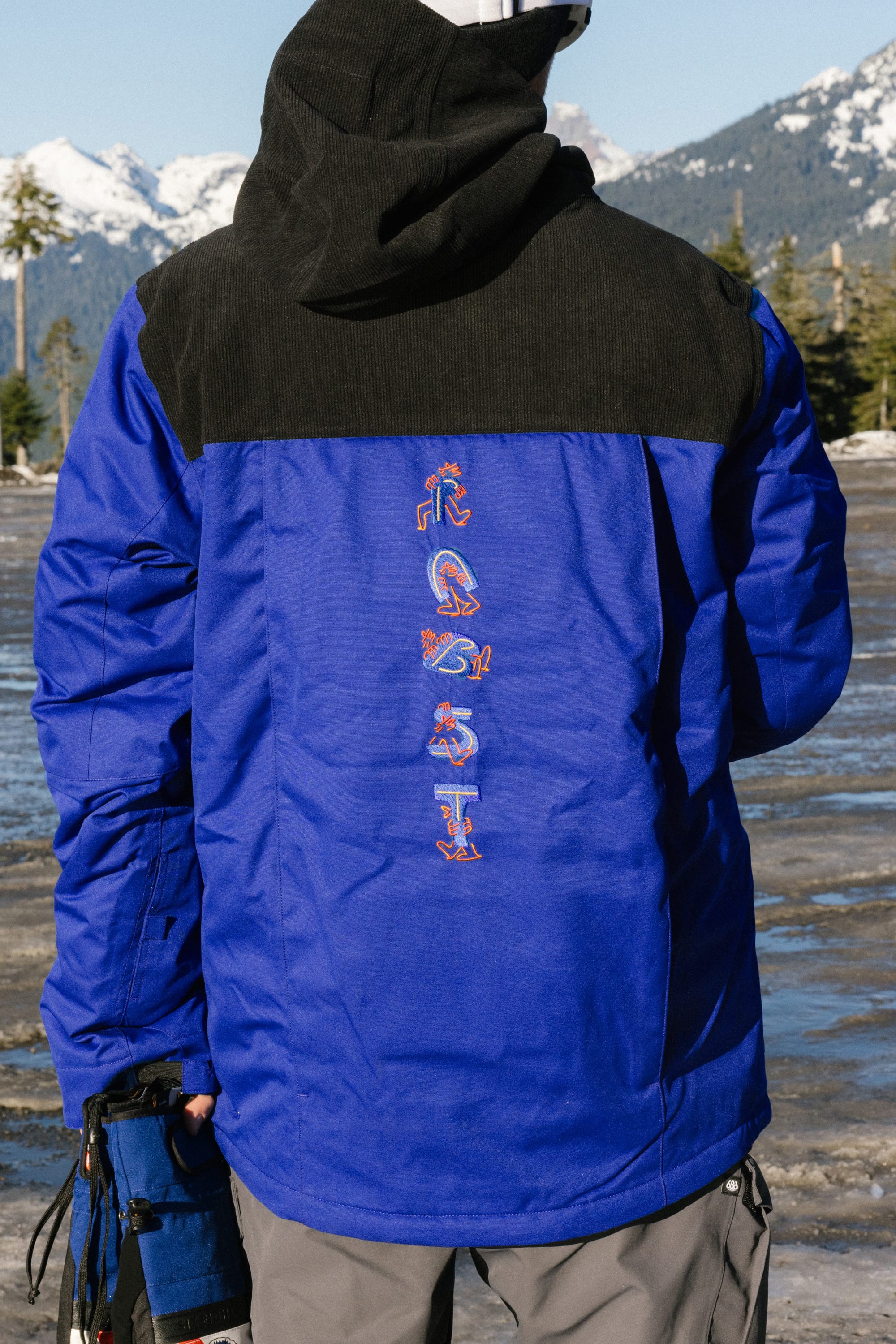 A person wearing the 686 Mens PBR Cooler Insulated Jacket stands outdoors on a snowy mountain landscape. The back of the blue and black 686 jacket features colorful stylized letters.
