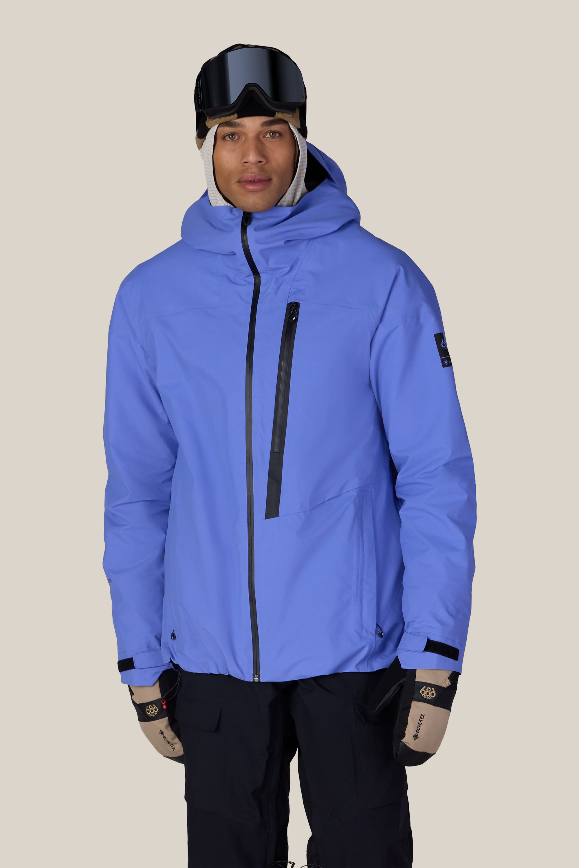 A person wearing the 686 Men’s GORE-TEX® GT Thermagraph® Jacket in light blue, along with beige gloves, black ski goggles, and dark pants stands against a plain beige background.