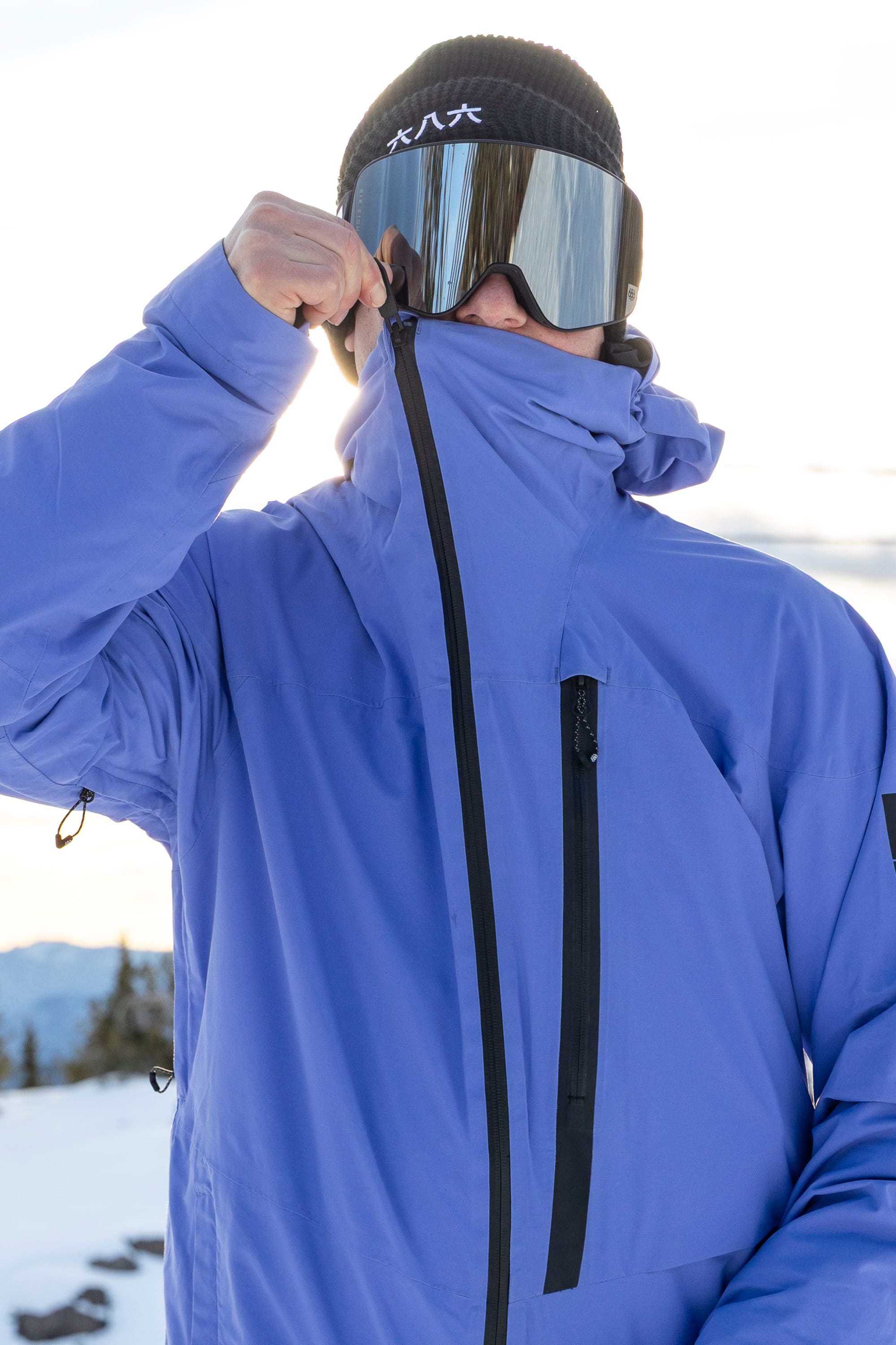 Wearing the 686 Men’s GORE-TEX® GT Thermagraph® Jacket, a person zips it up over their face. They have on reflective ski goggles and a black beanie with white text, standing outdoors in a snowy mountain setting.