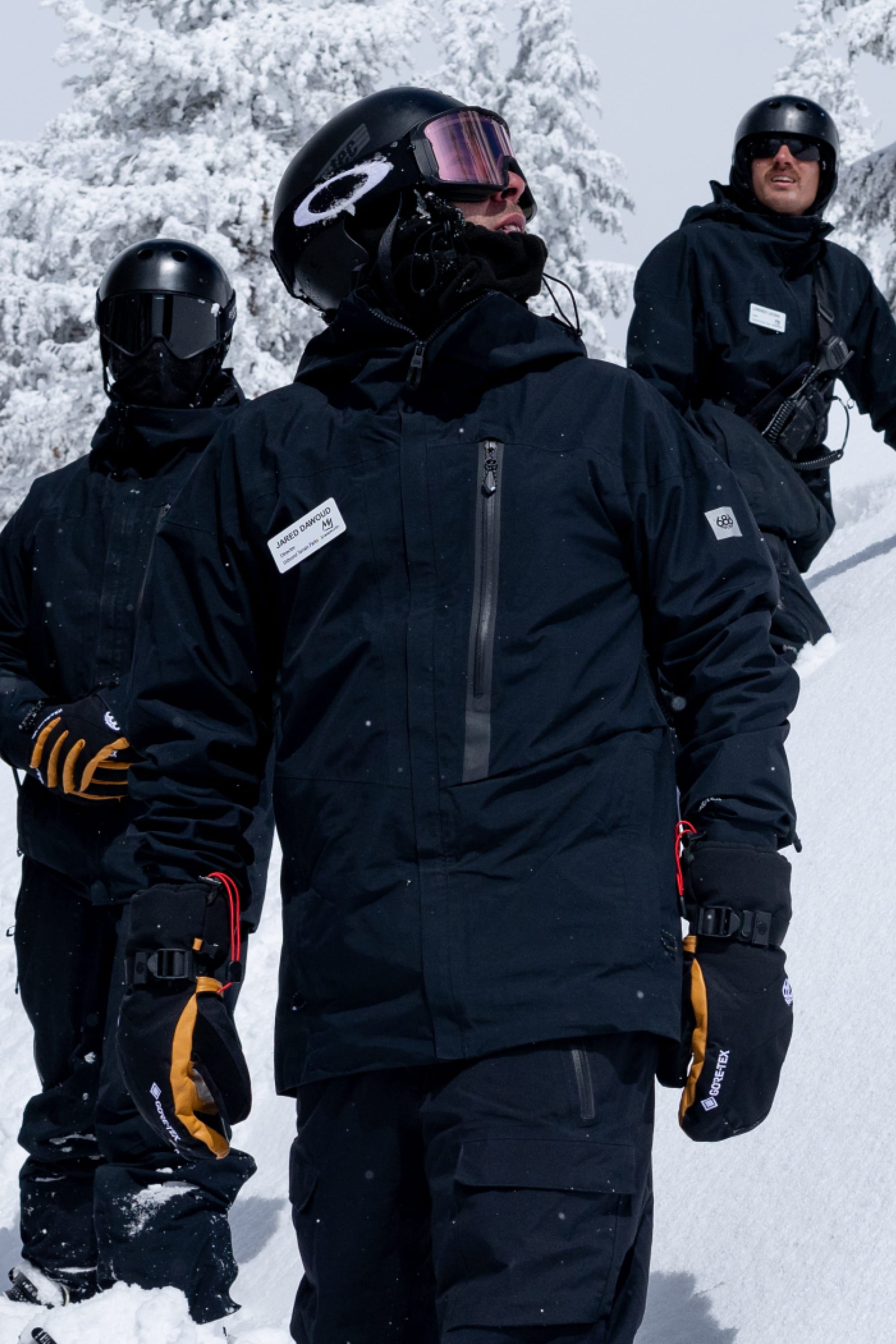 Three people wearing 686 Men's GORE-TEX GT Thermagraph® Jackets by 686, along with helmets and goggles, stand in a snowy landscape and look upward, with snow-covered trees in the background.