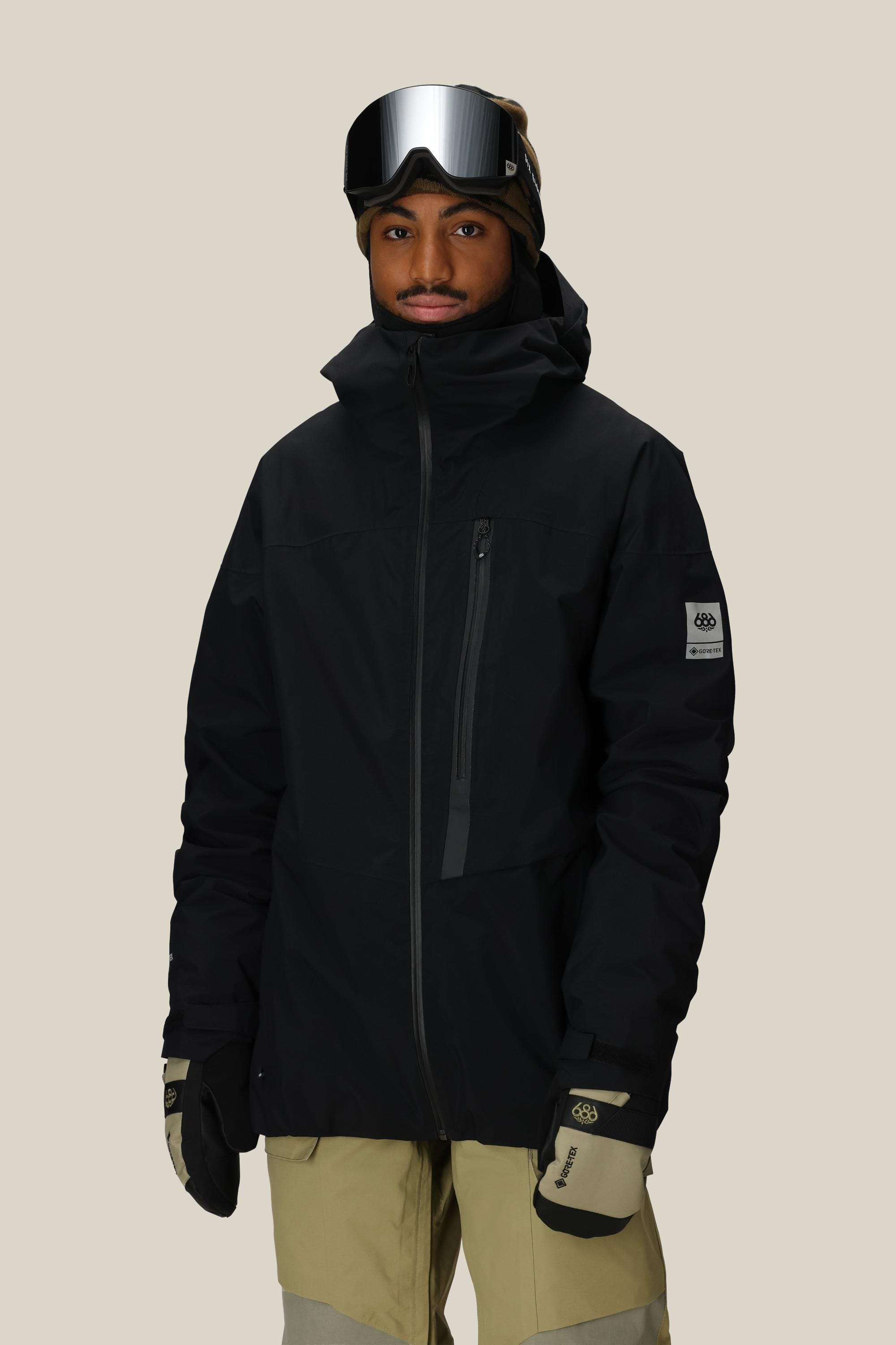 A person wearing the 686 Men’s GORE-TEX® GT Thermagraph® Jacket stands facing the camera against a plain background, paired with tan snow pants, tan and black gloves, and ski goggles.