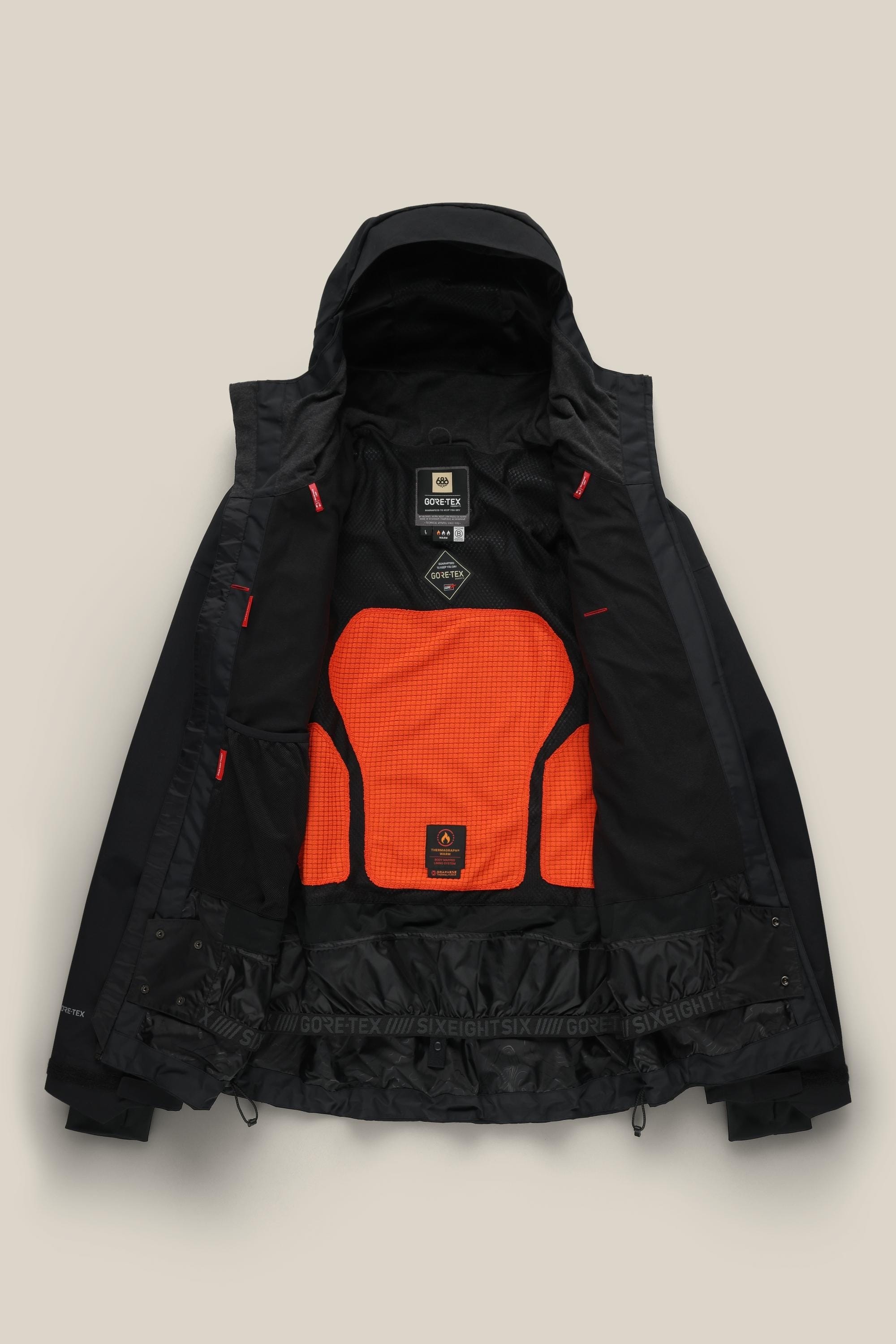 A 686 Men’s GORE-TEX® GT Thermagraph® Jacket in black is shown open on a beige background, revealing an orange mesh inner panel, black lining, interior labels, and red accents inside the jacket.