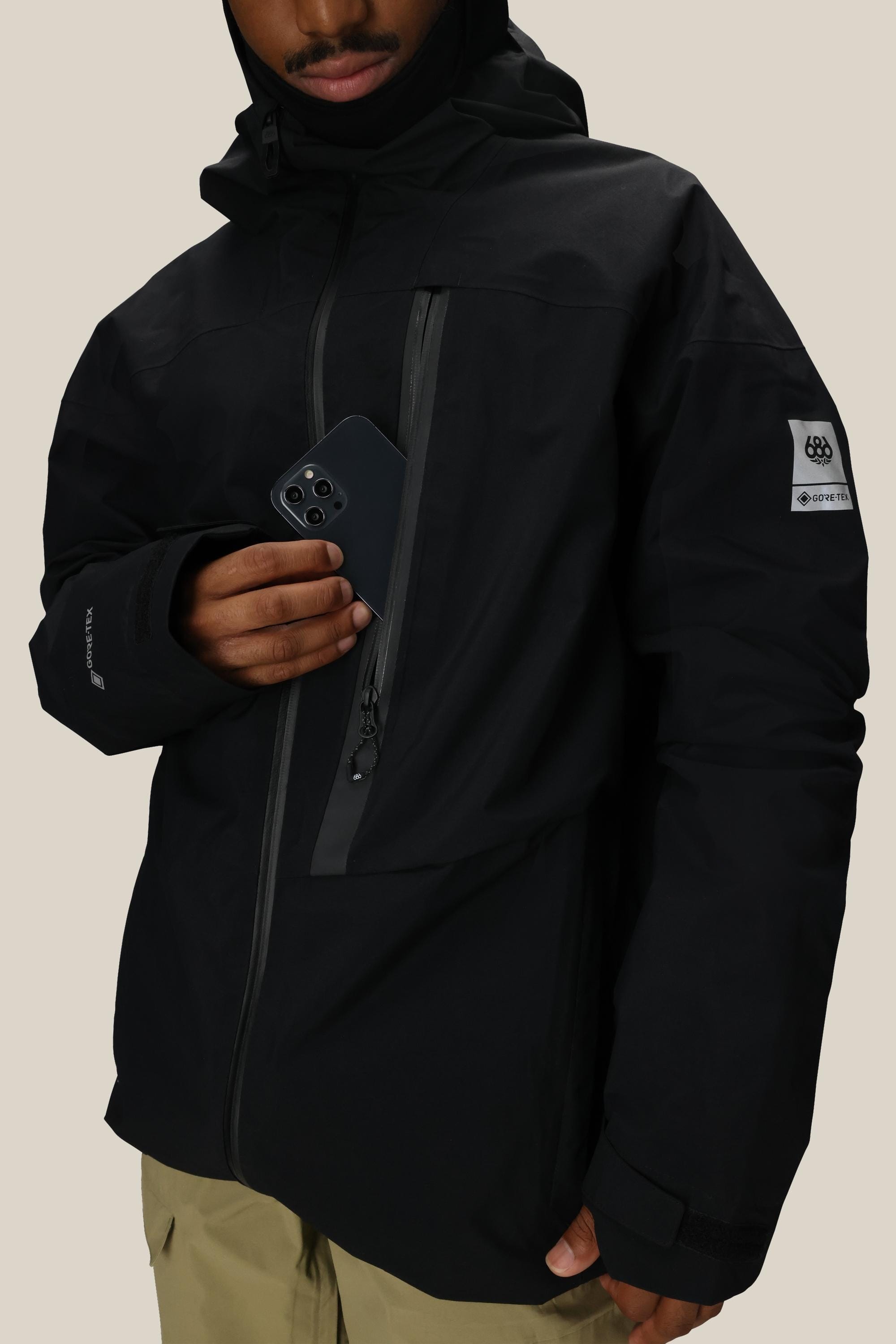 A person wearing the 686 Men’s GORE-TEX® GT Thermagraph® Jacket in black slides a smartphone into the chest pocket. The jacket, featuring a sleeve badge and made by 686, is paired with light-colored pants against a plain beige background.