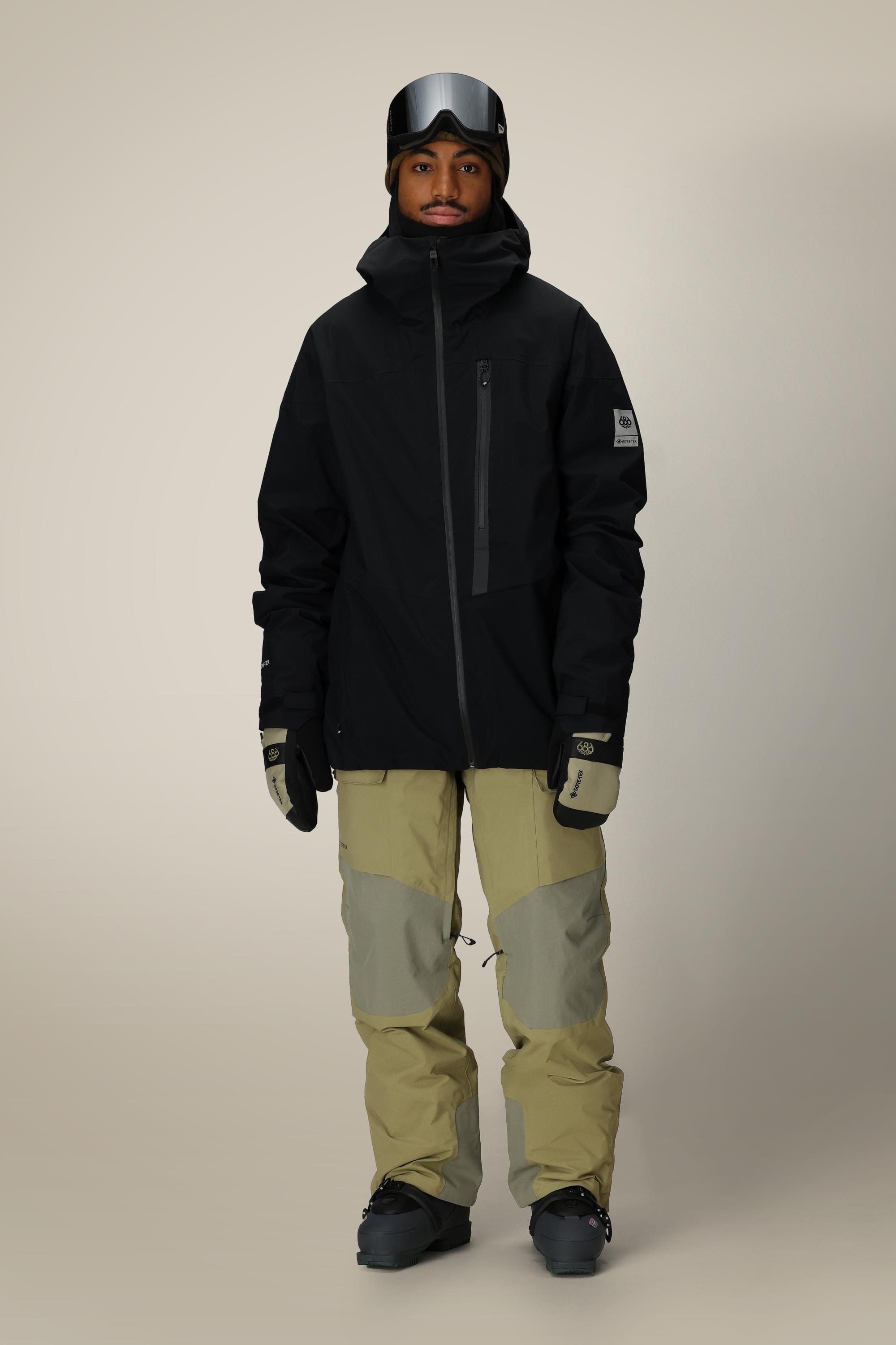 A person wearing the 686 Men’s GORE-TEX® GT Thermagraph® Jacket stands facing forward against a plain light background, paired with tan and gray snow pants, gloves, ski goggles, and boots.