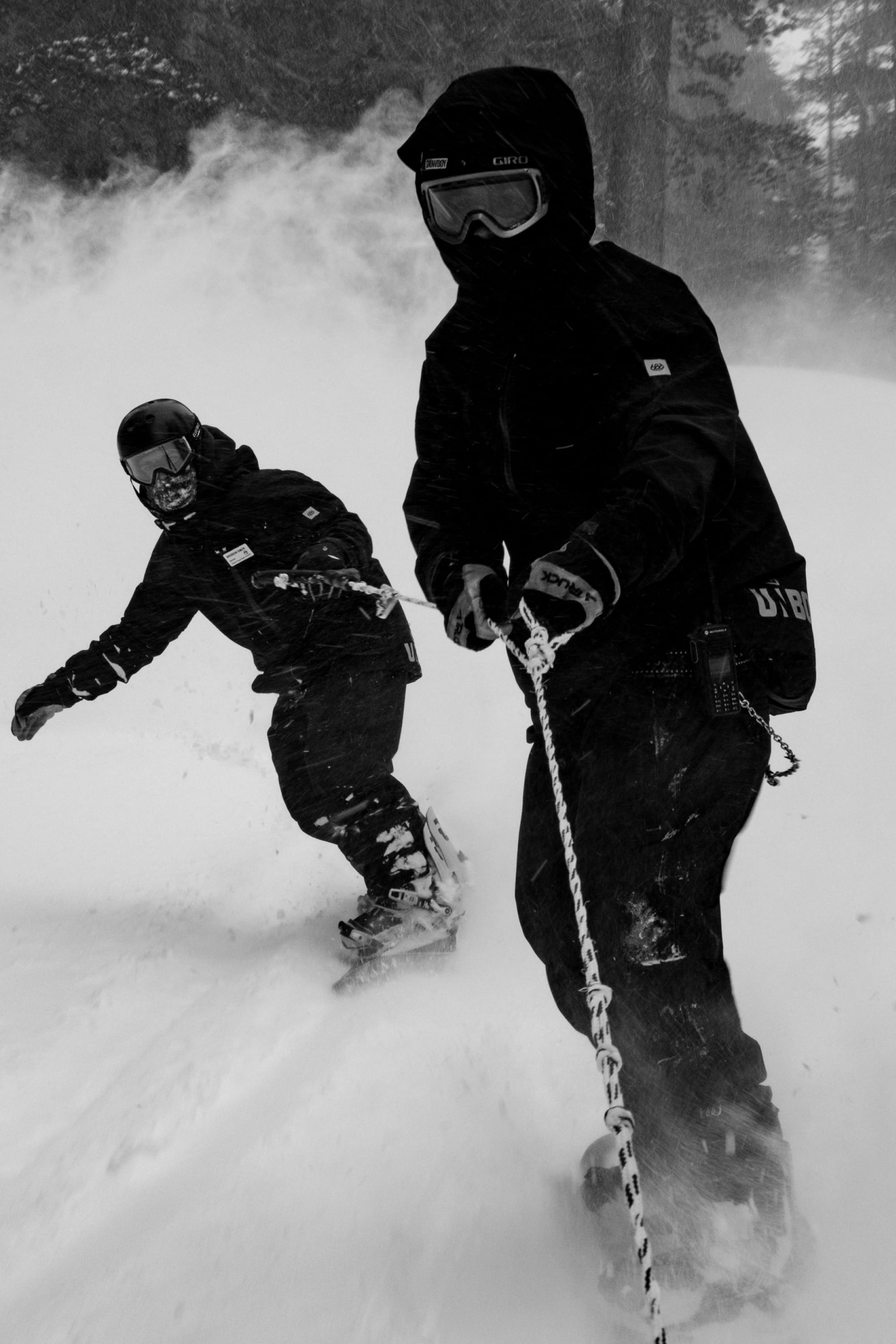 Two people in winter gear—one wearing the 686 Men’s GORE-TEX® GT Thermagraph® Jacket—snowboard down a snowy slope, with one holding a rope attached to the other. Snow swirls around them as trees stand in the background, all captured in black and white.