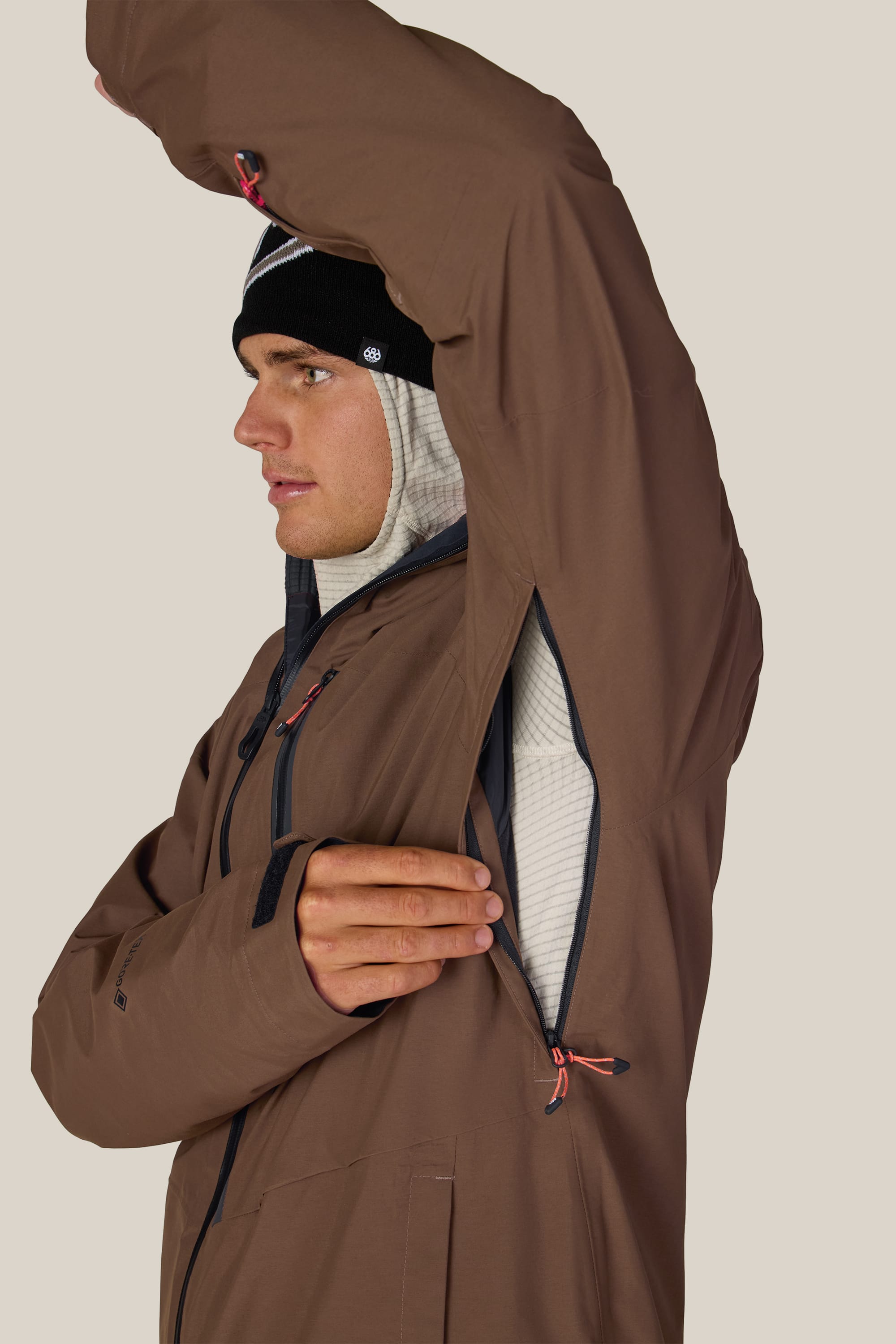 A person wearing the 686 Men’s GORE-TEX® GT Thermagraph® Jacket in brown with a black beanie unzips the large underarm ventilation zipper, revealing a light gray inner layer. The background is plain and beige.