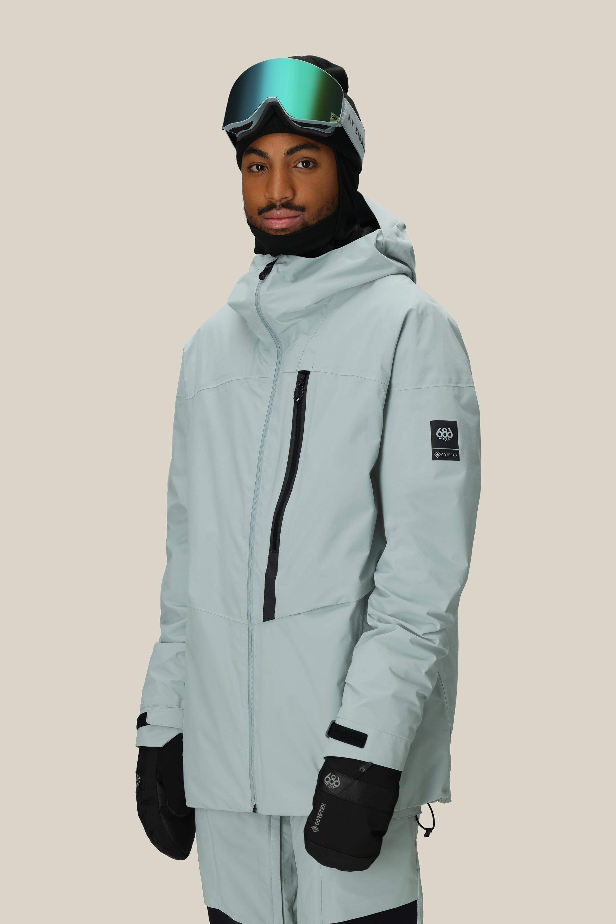 A person wears the 686 Men's GORE-TEX GT Thermagraph® Jacket, black gloves, a black beanie, and reflective ski goggles while standing against a plain beige background.