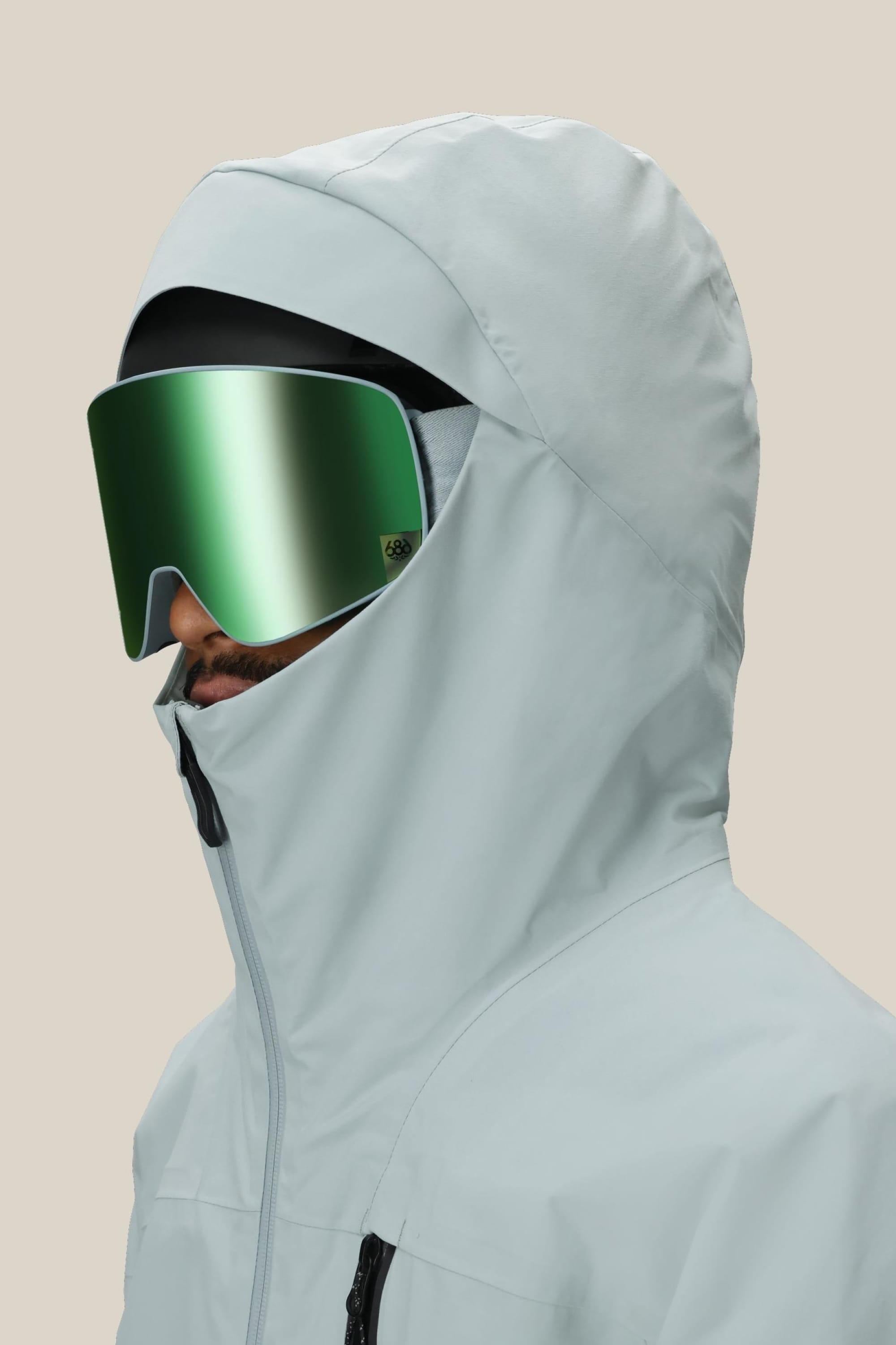 A person wearing the 686 Men's GORE-TEX GT Thermagraph® Jacket in light gray and large reflective green ski goggles faces left against a plain beige background.