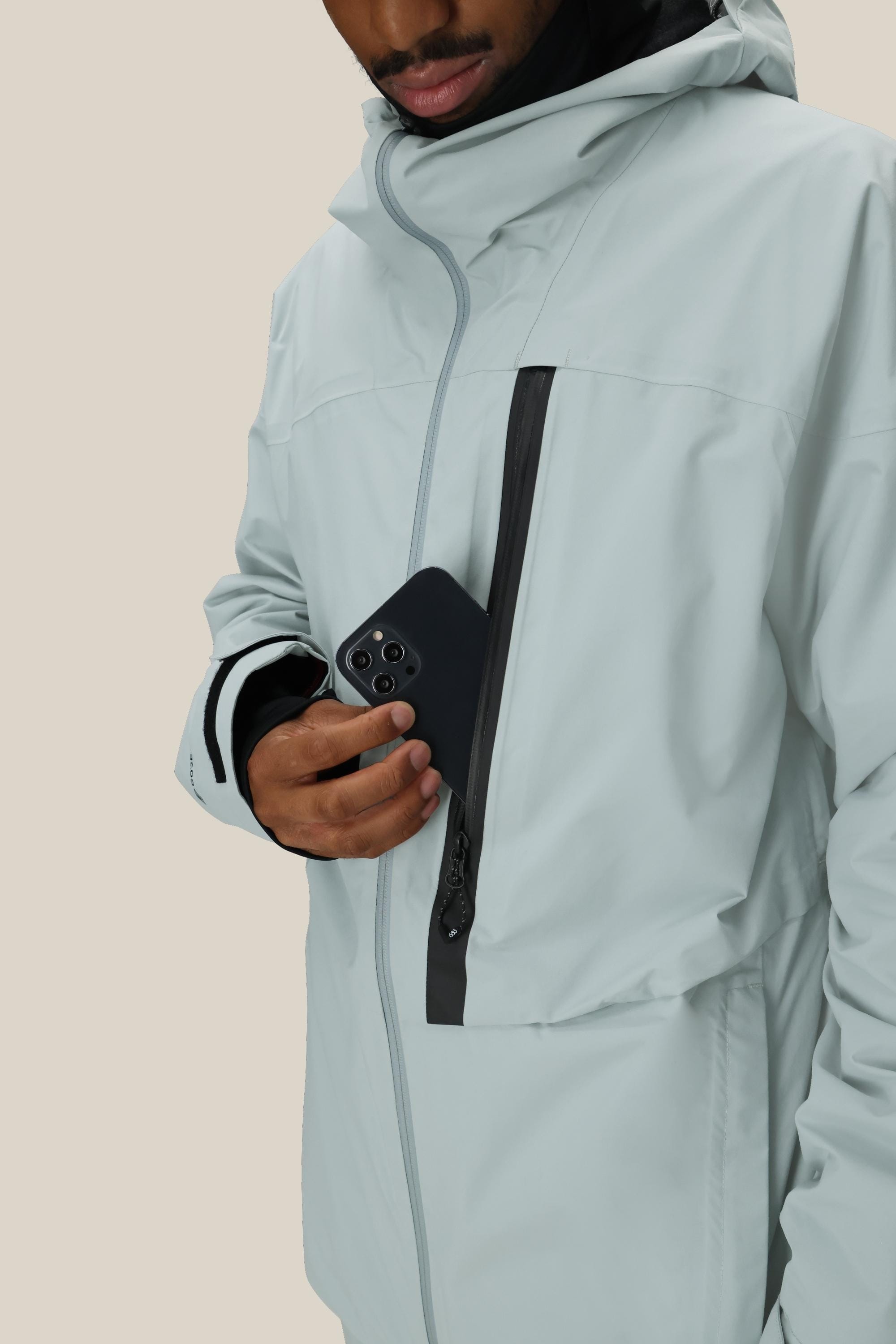 A person wearing the 686 Men's GORE-TEX GT Thermagraph® Jacket places a smartphone into its zippered chest pocket. The light gray jacket features black zipper details, and the person's face is partially visible.