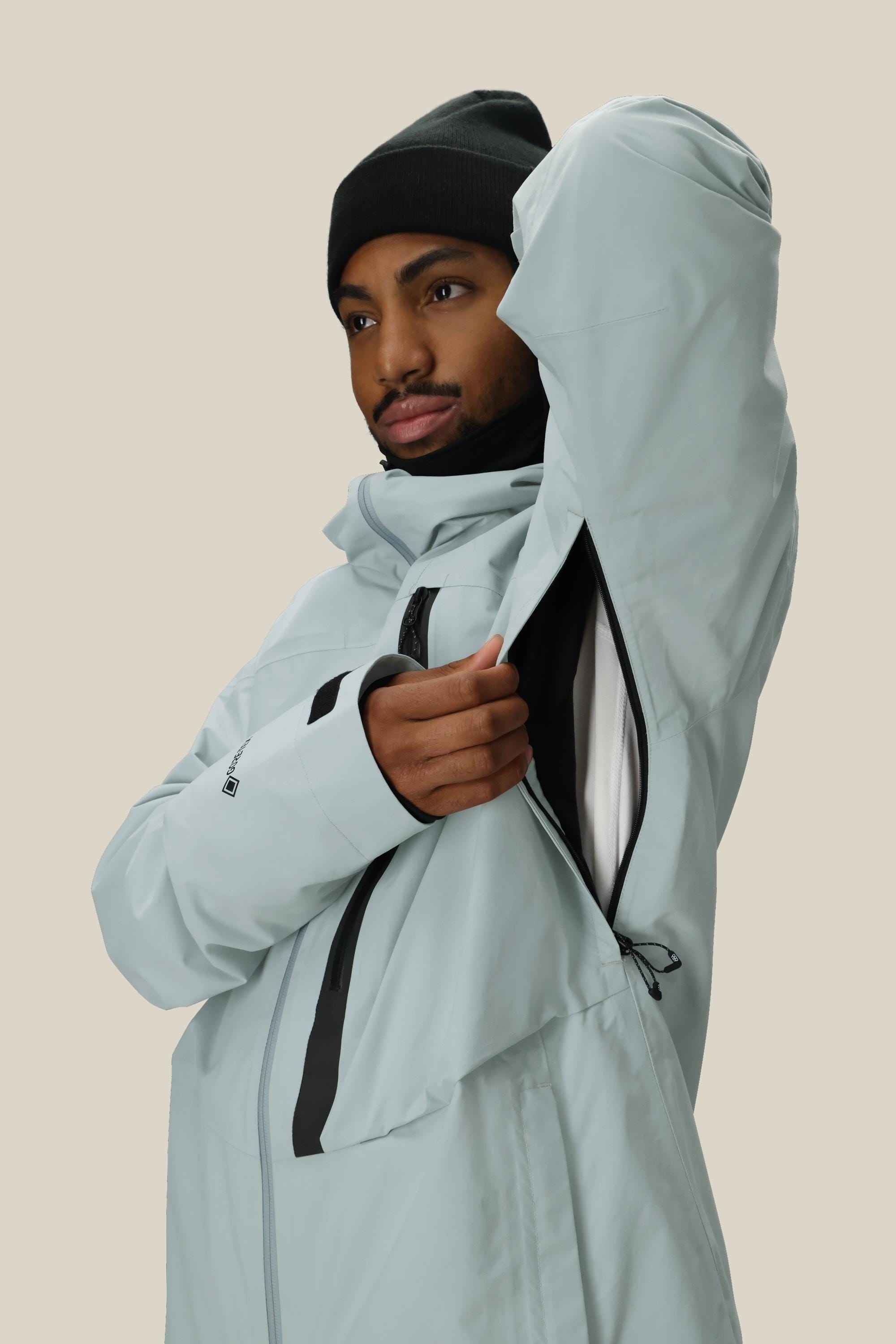 A person wearing the 686 Men's GORE-TEX GT Thermagraph® Jacket in light gray and a black beanie unzips an underarm vent, showing the jacket’s inner lining against a neutral background.