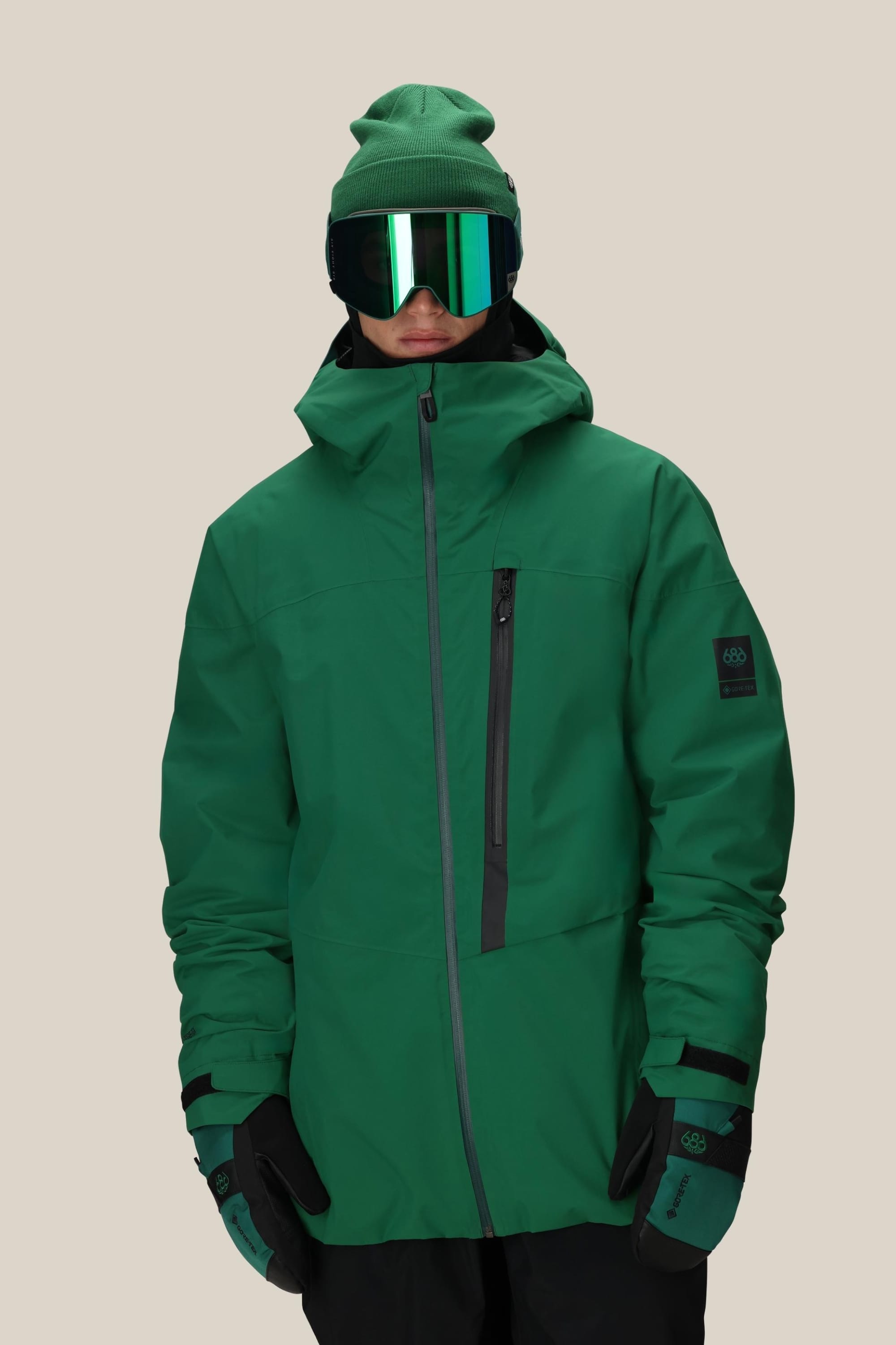 A person wearing the 686 Men's GORE-TEX GT Thermagraph® Jacket, green gloves, a green beanie, and reflective ski goggles stands against a plain beige background.
