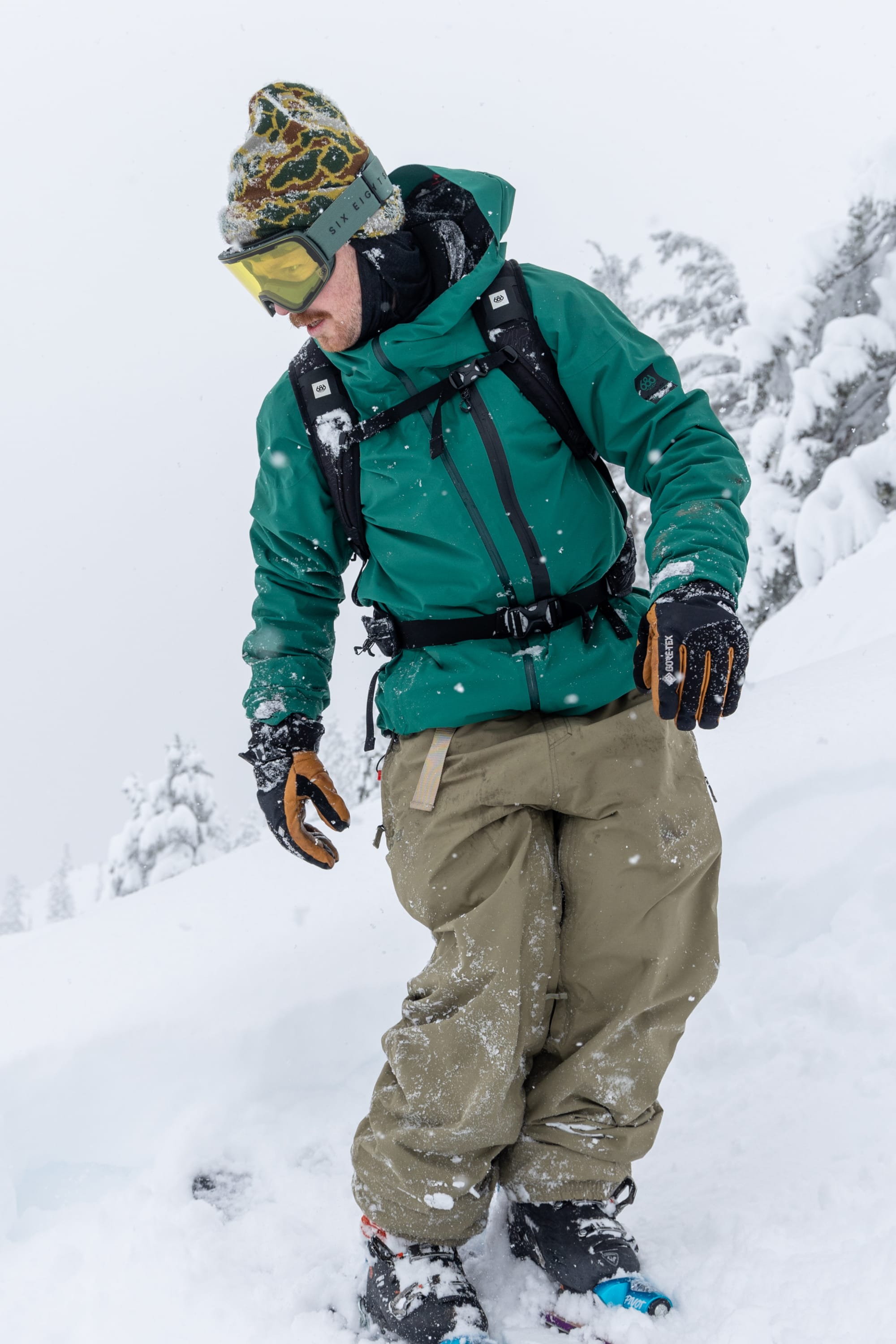Wearing the 686 Men's GORE-TEX GT Thermagraph® Jacket, a person stands on a snowy slope among snow-covered trees, dressed in winter gear with tan pants, gloves, goggles, and a beanie.