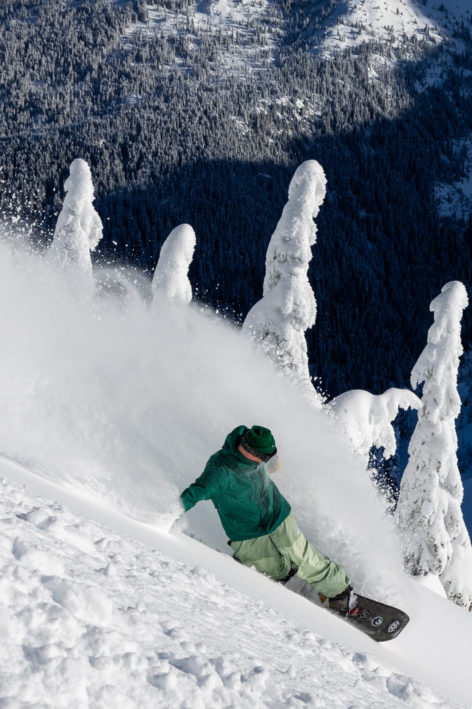 Wearing the 686 Men's GORE-TEX GT Thermagraph® Jacket, a snowboarder carves through deep powder on a snowy mountain slope, with snow-covered trees and a forested valley in the background.