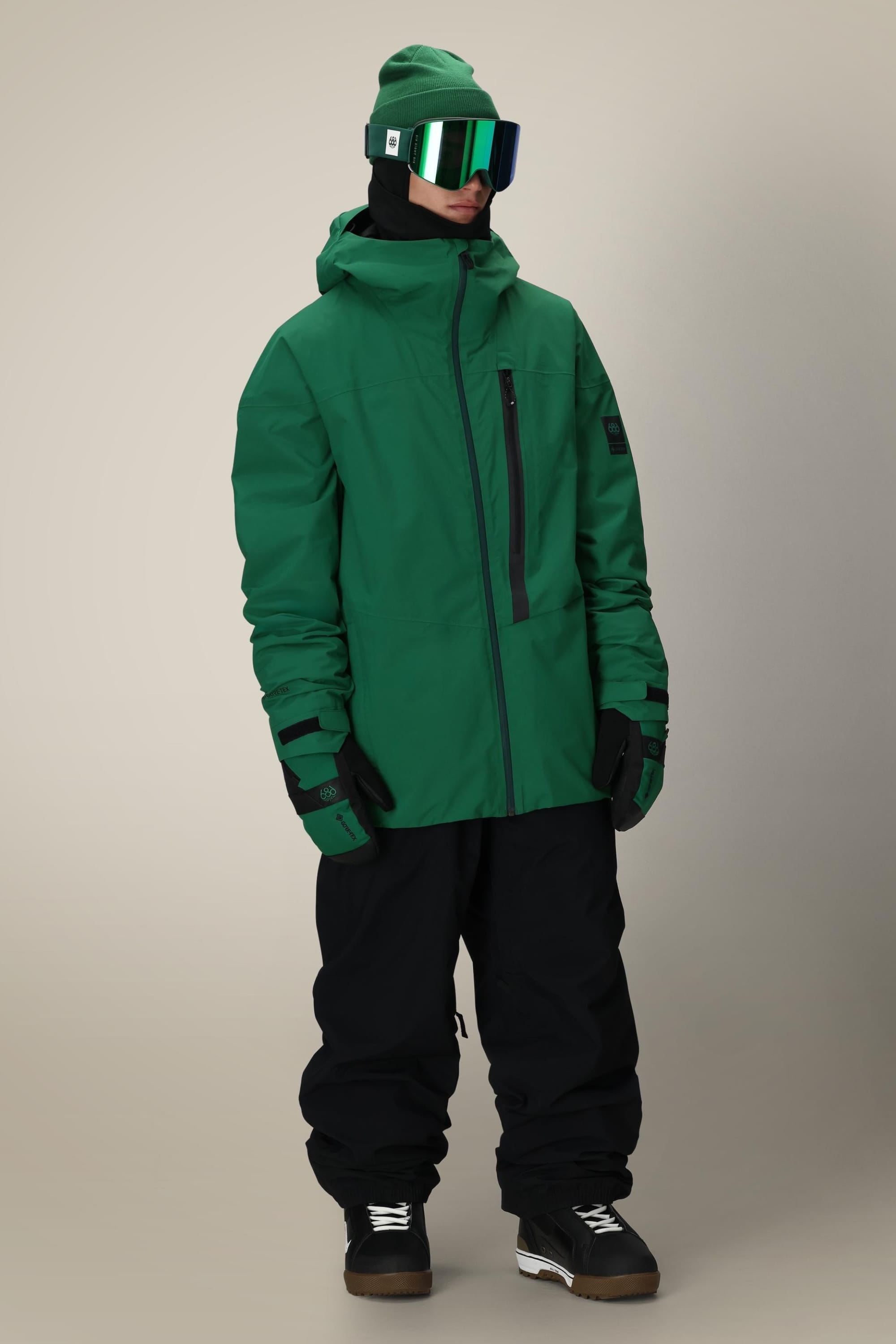 A person wearing the 686 Men's GORE-TEX GT Thermagraph® Jacket, green gloves, beanie, and ski goggles—paired with black snow pants and boots—stands facing slightly left against a neutral background.