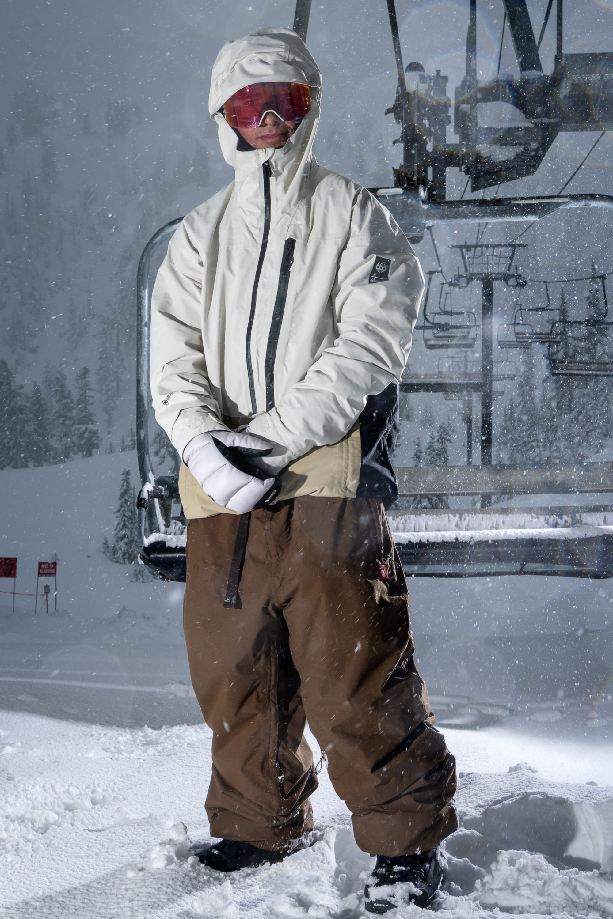 A person in winter gear stands in the snow near a ski lift, wearing the 686 Men’s GORE-TEX® GT Thermagraph® Jacket, brown pants, gloves, and red goggles as snow falls and trees line the snowy background.