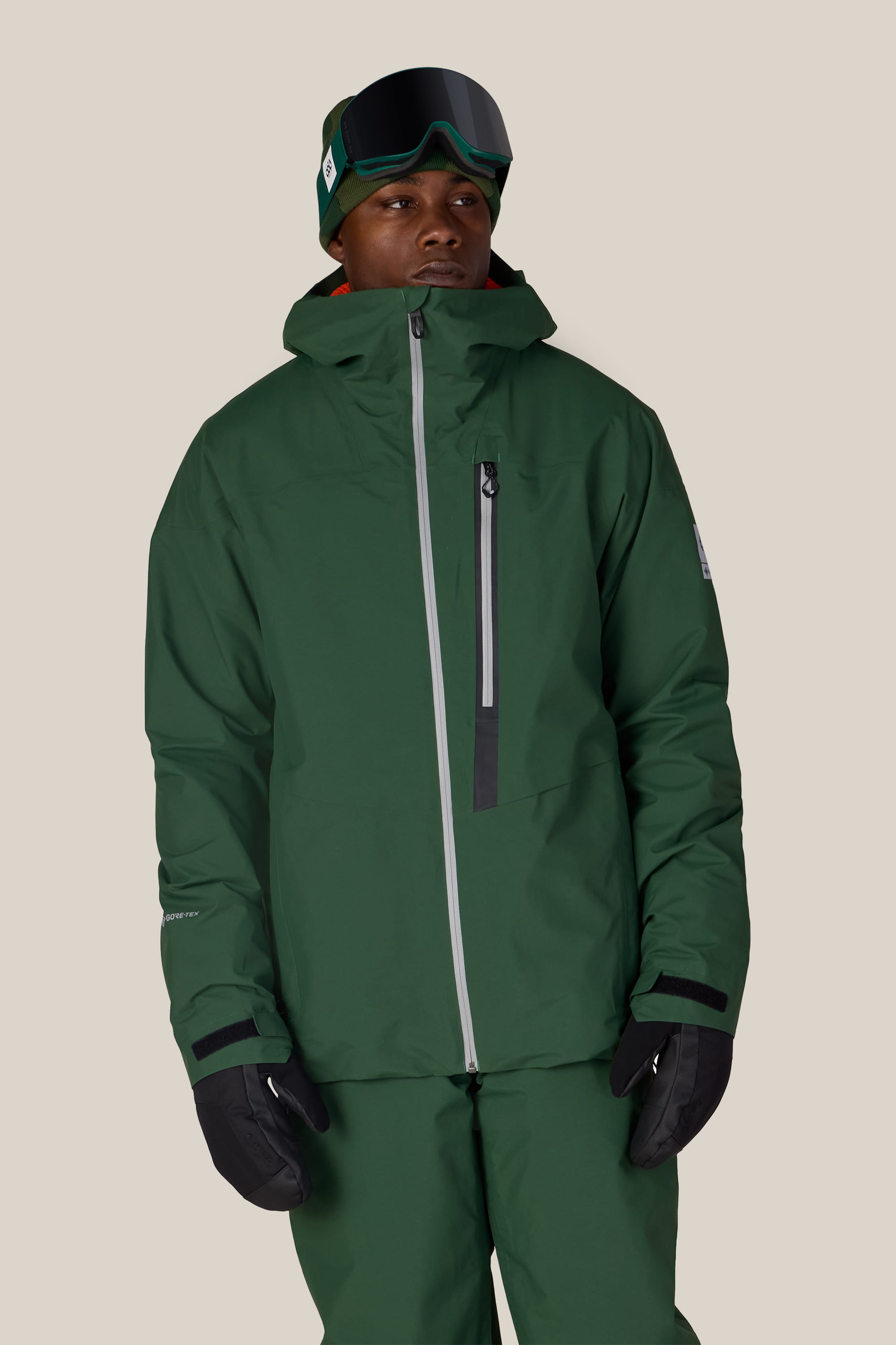 A person wearing the 686 Men’s GORE-TEX® GT Thermagraph® Jacket by 686, paired with matching green pants, black gloves, and ski goggles stands against a plain light background.