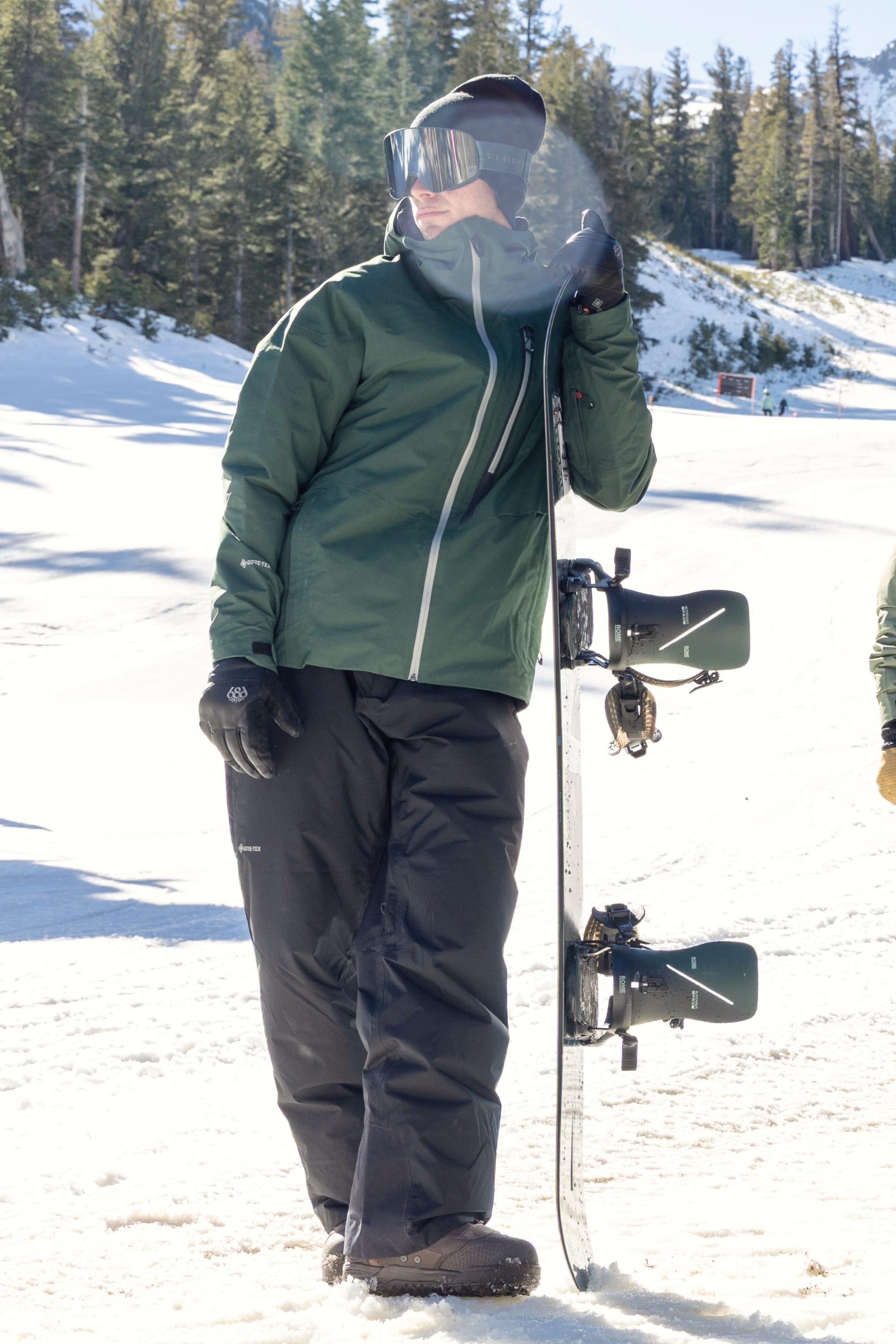 A person in winter gear stands on snowy ground, leaning on a snowboard. They wear the 686 Men’s GORE-TEX® GT Thermagraph® Jacket by 686, along with black snow pants, gloves, a black beanie, and ski goggles, with trees and sunlight in the background.