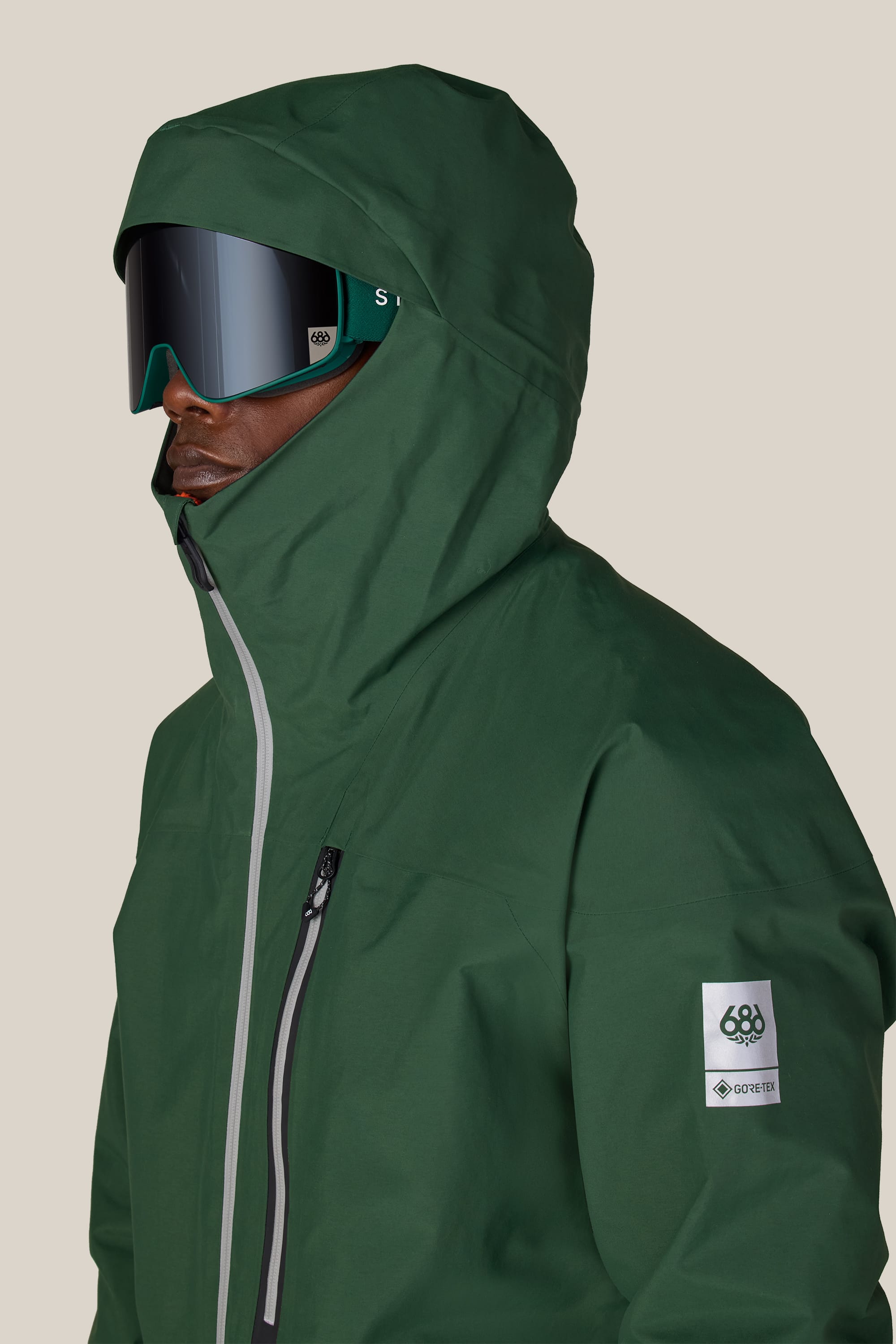 A person wearing the 686 Men’s GORE-TEX® GT Thermagraph® Jacket in green, featuring a visible GORE-TEX patch, along with black ski goggles, is facing left against a plain light background.