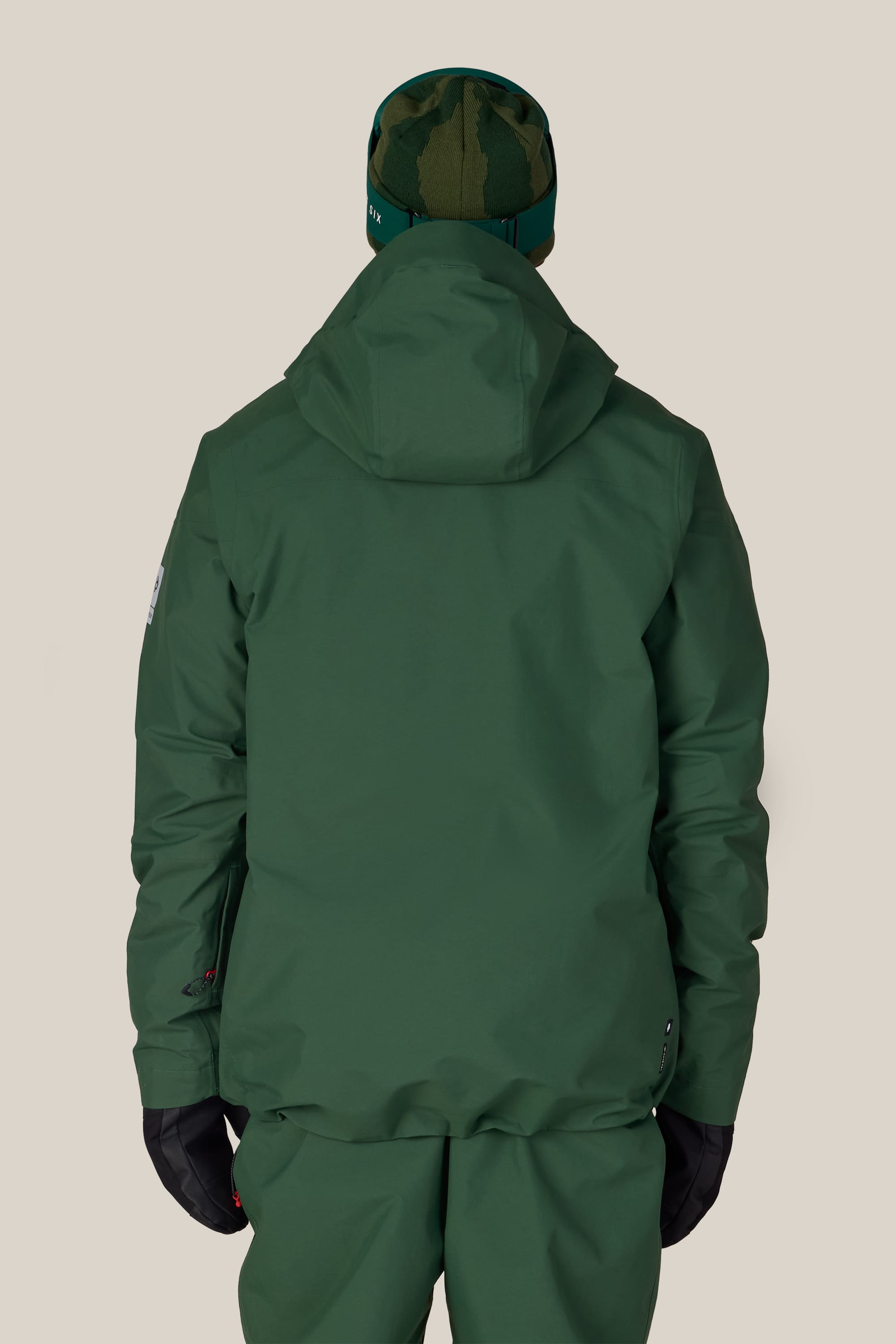 A person wearing the 686 Men’s GORE-TEX® GT Thermagraph® Jacket by 686 is shown from the back against a plain beige background, styled with matching green pants, black gloves, and a green patterned beanie.