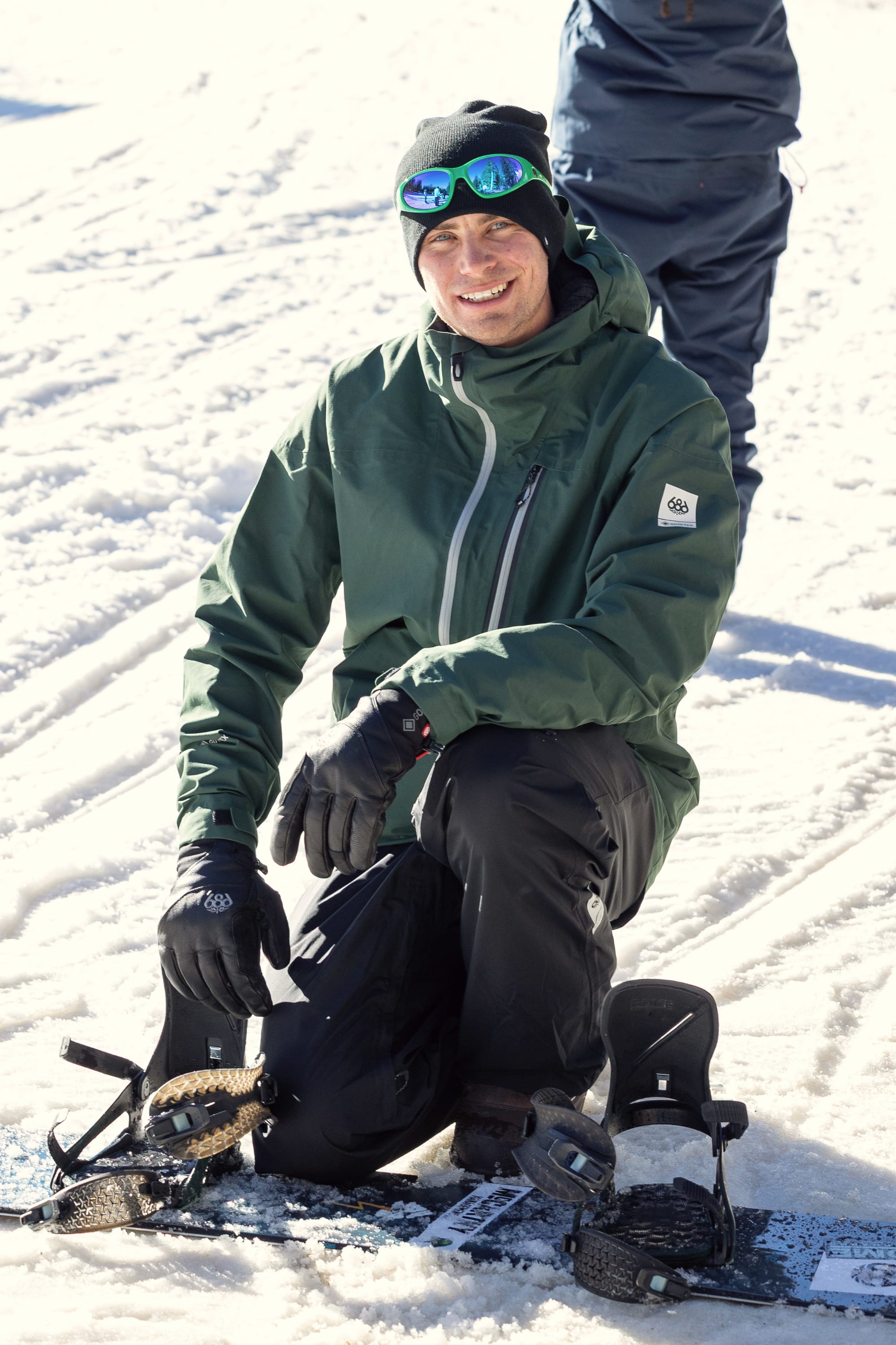 A man wearing the 686 Men’s GORE-TEX® GT Thermagraph® Jacket by 686, along with black gloves and sunglasses, kneels on snow next to a snowboard and smiles at the camera. Another person’s legs are visible in the background.