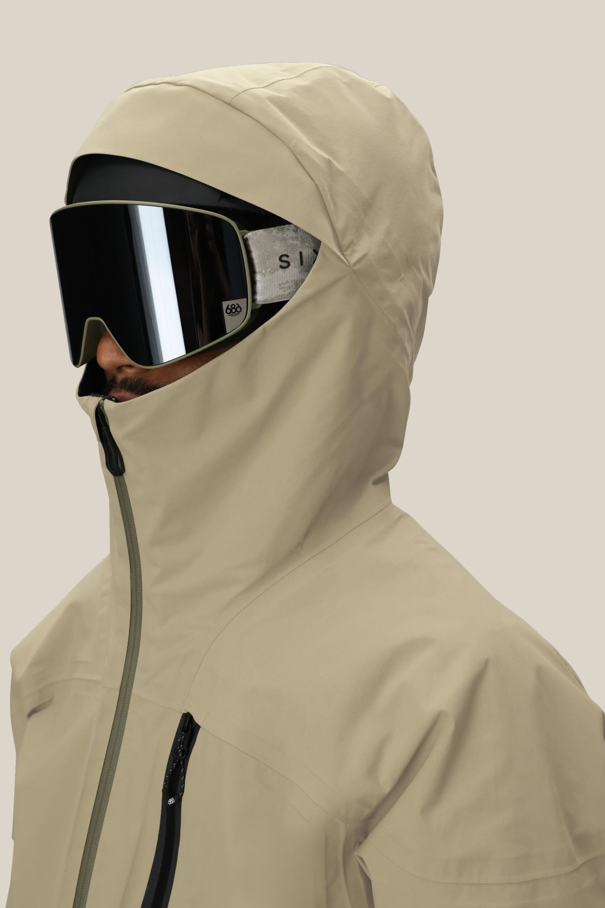 A person wears the 686 Men's GORE-TEX GT Thermagraph® Jacket, hood up and zipped to cover most of their face, along with large black ski goggles, against a plain beige background.
