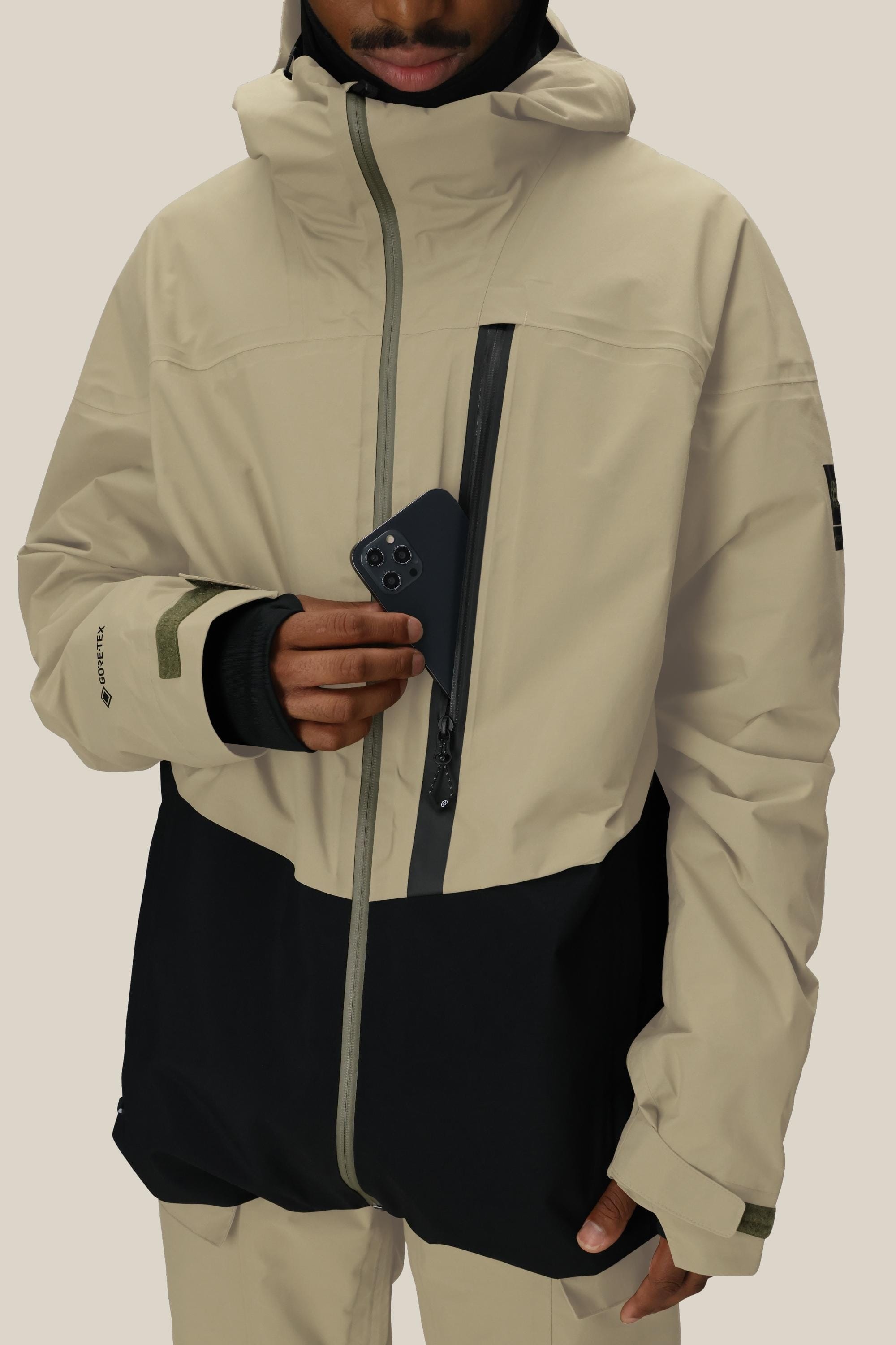 A person wearing the 686 Men's GORE-TEX GT Thermagraph® Jacket in beige and black places a black smartphone into its vertical zippered chest pocket. The 686 jacket features waterproof nylon, a high collar, and partially reveals the wearer’s face.