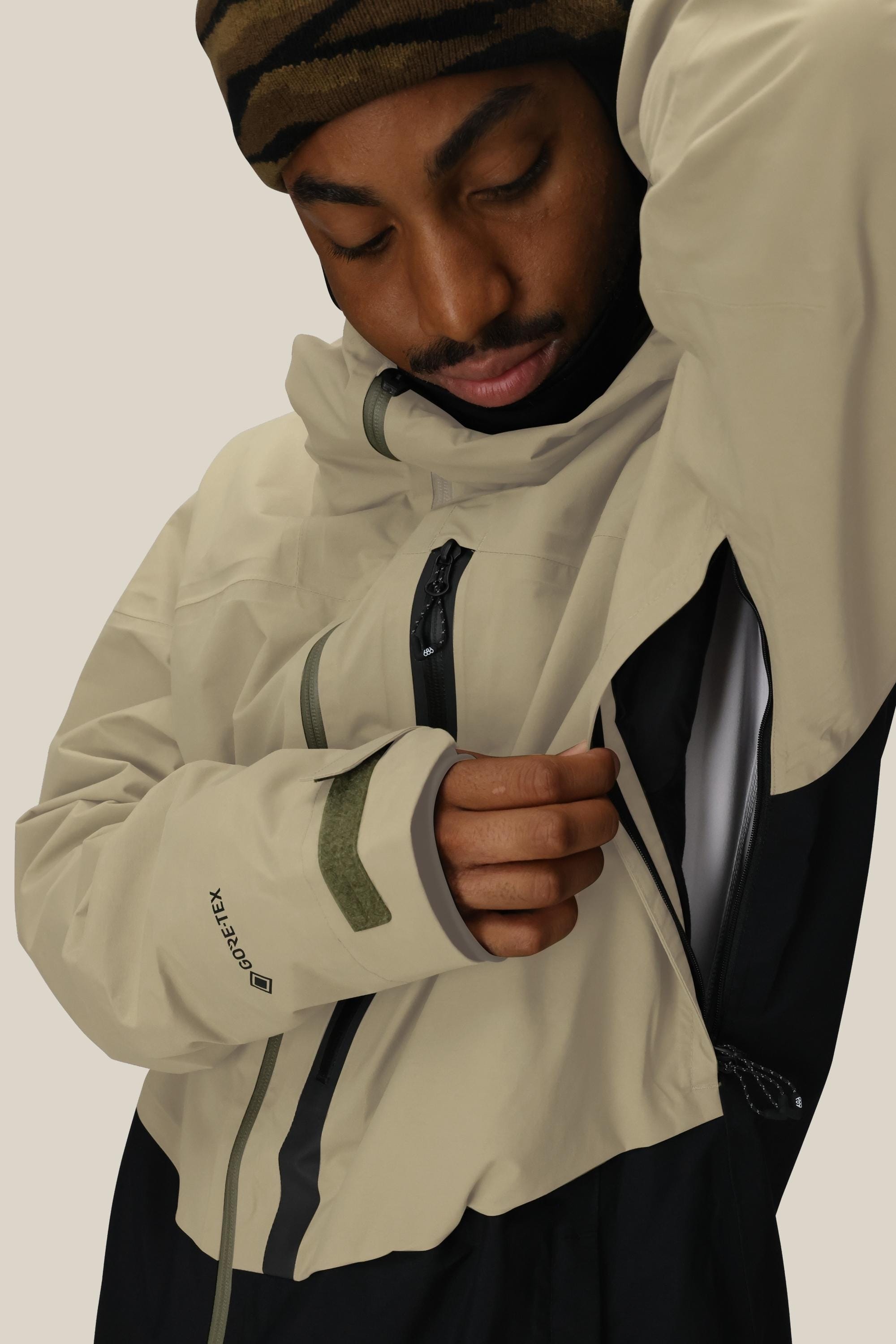 A man in a 686 Men's GORE-TEX GT Thermagraph® Jacket—beige and black with green accents—pairs it with a brown patterned beanie and opens the underarm zipper for ventilation; the jacket’s curved cuff adds stylish detail.